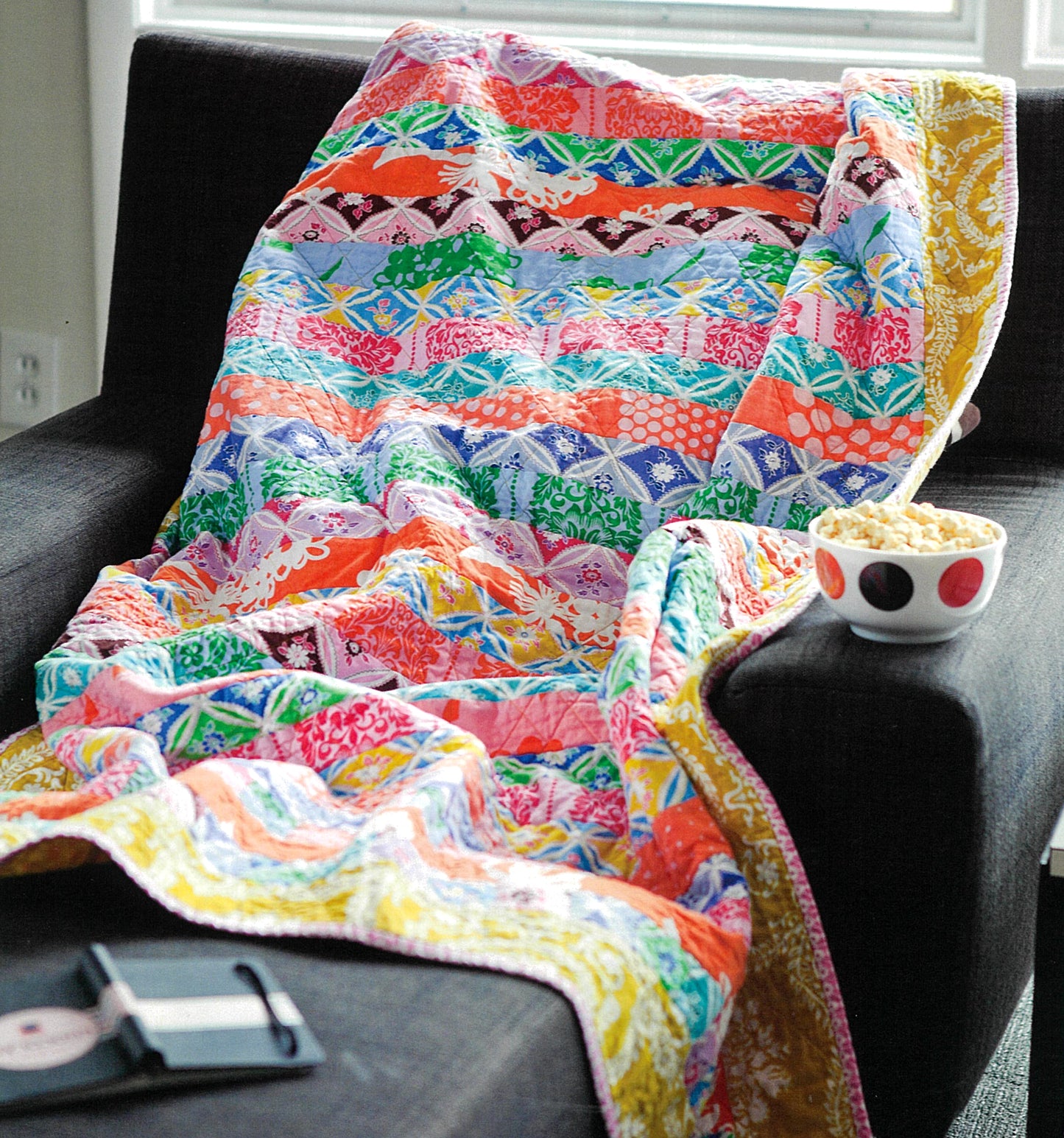 Leisure Arts’ "The Best of Quilty" patchwork quilt, featuring vibrant patterns inspired by beginner embroidery and decorative stitches, is draped over a dark armchair beside a bowl of popcorn and smartphone. Sunlight streams through the window behind.