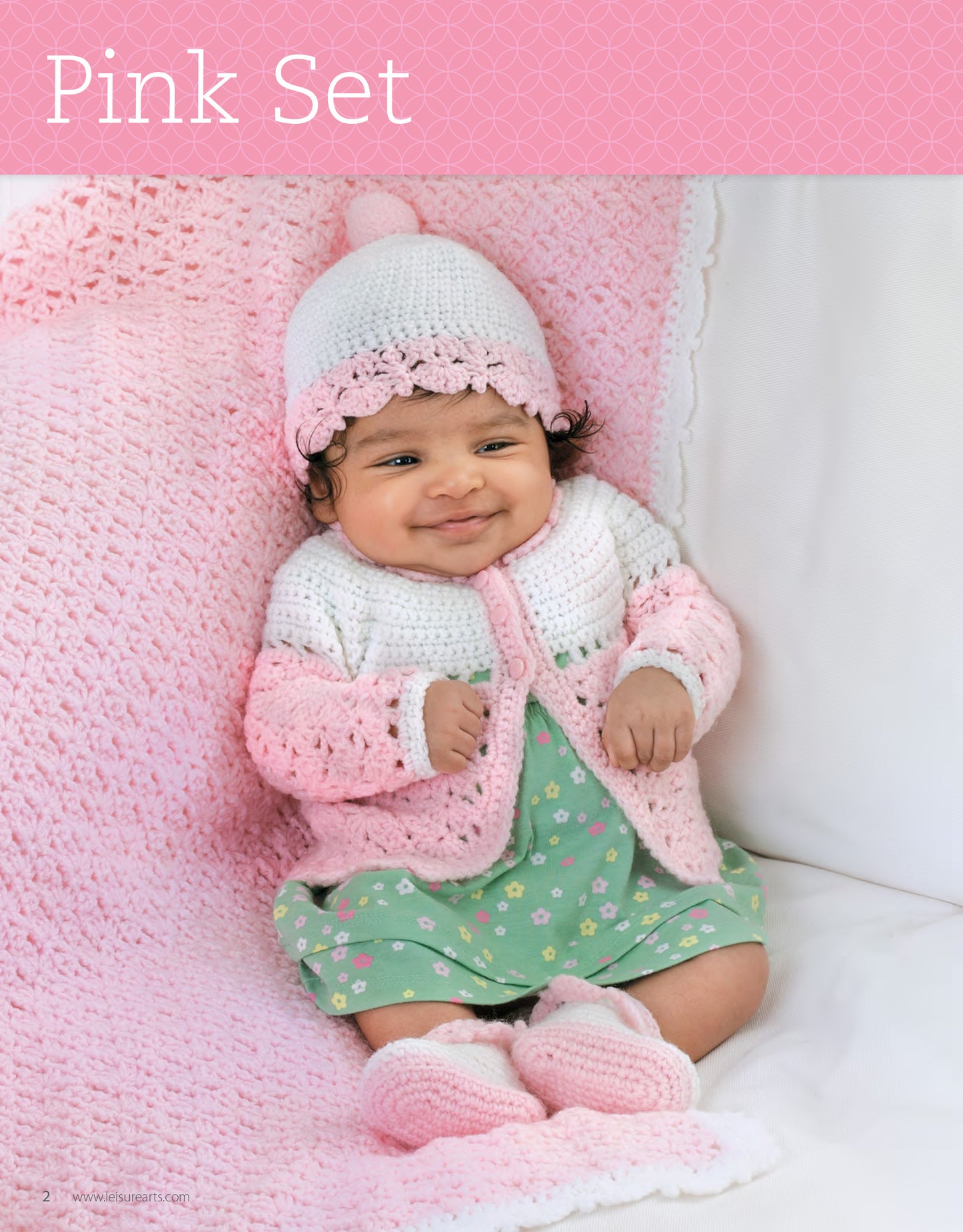 A smiling baby in a green floral dress sits on a pink crocheted blanket, wearing the My Very First Layette 2 set by Leisure Arts—a pink and white crocheted hat, cardigan, and booties.