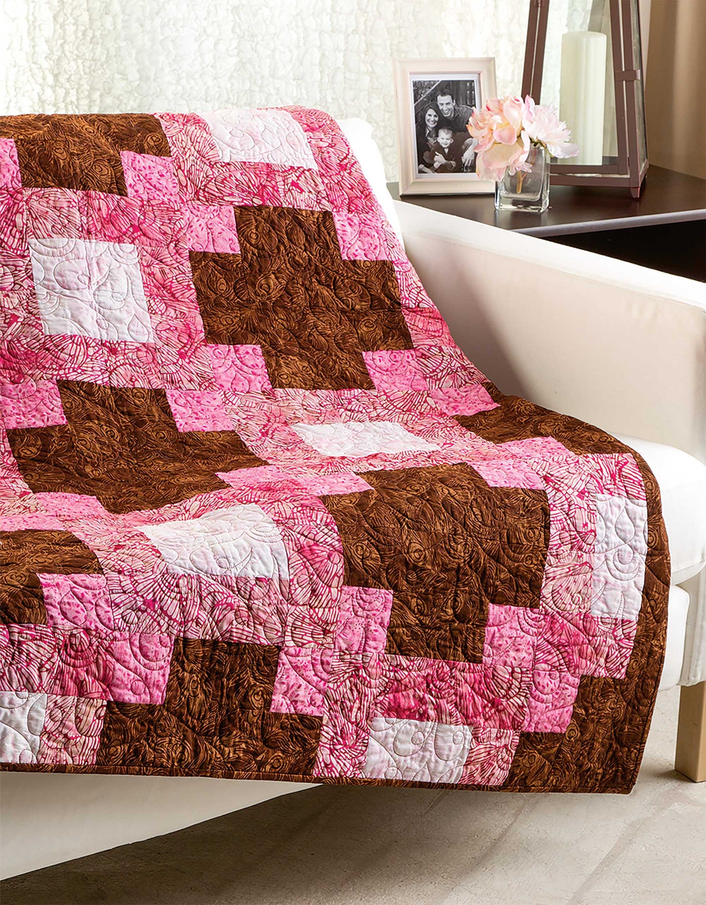 The Quick & Easy Quilts book by Leisure Arts is shown with a pink, brown, and white patchwork quilt draped over a white sofa; behind it, a side table displays pink flowers, a black-and-white photo, lamp, and décor.