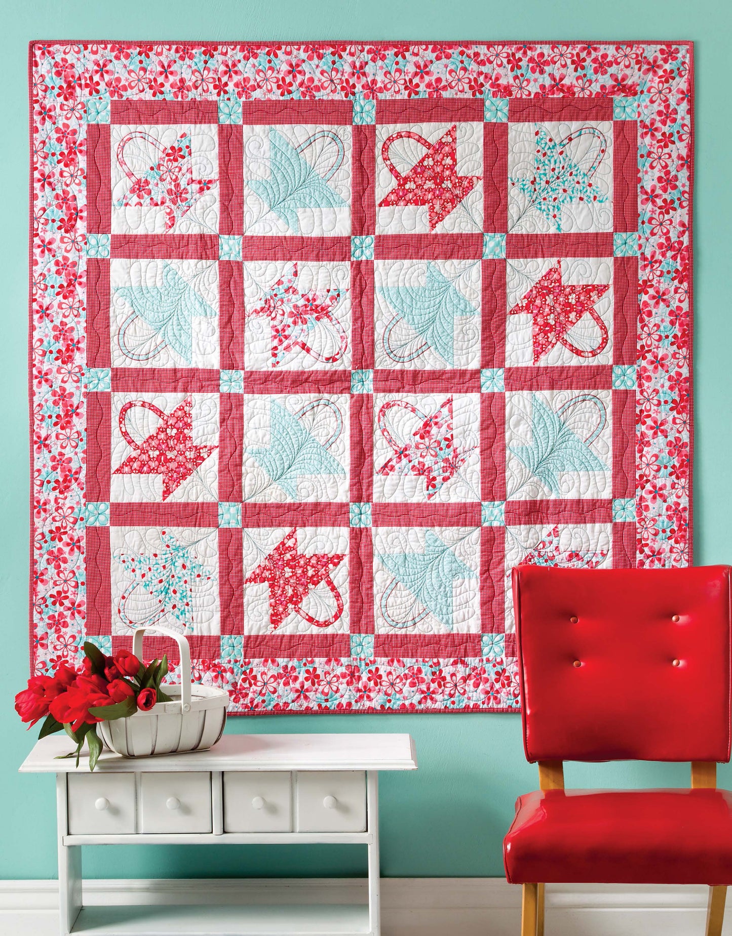 The Lap & Throw Quilts by Leisure Arts, featuring vibrant pink, red, and blue patchwork, is displayed on a teal wall above a white console table with a basket of red flowers and next to a shiny red chair.