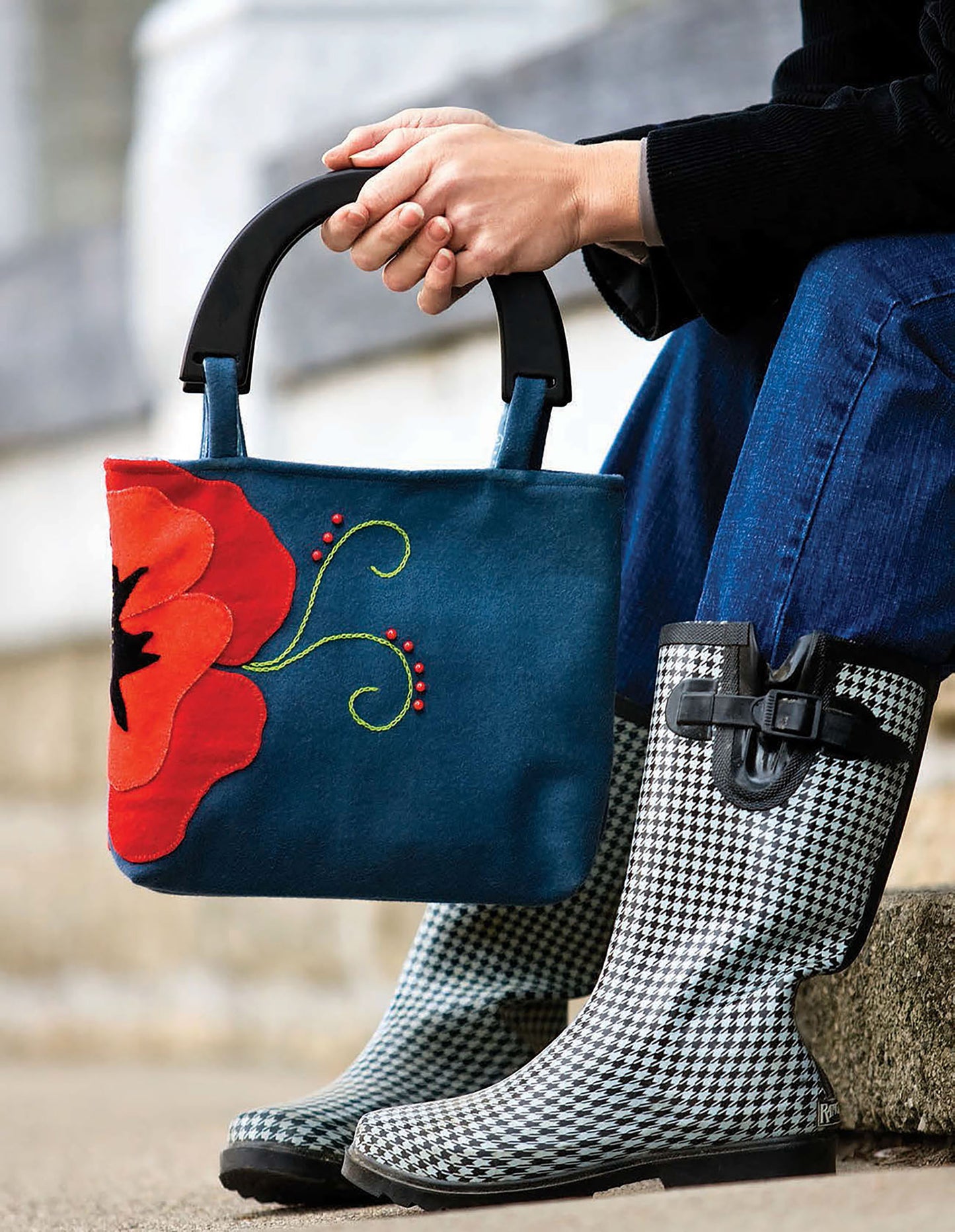 Wearing blue jeans and houndstooth rain boots, a person sits on steps holding a Leisure Arts Quilted Purses & Totes bag—a stylish blue handbag with a large red flower and green embroidery near the black handles, inspiring tote designs.