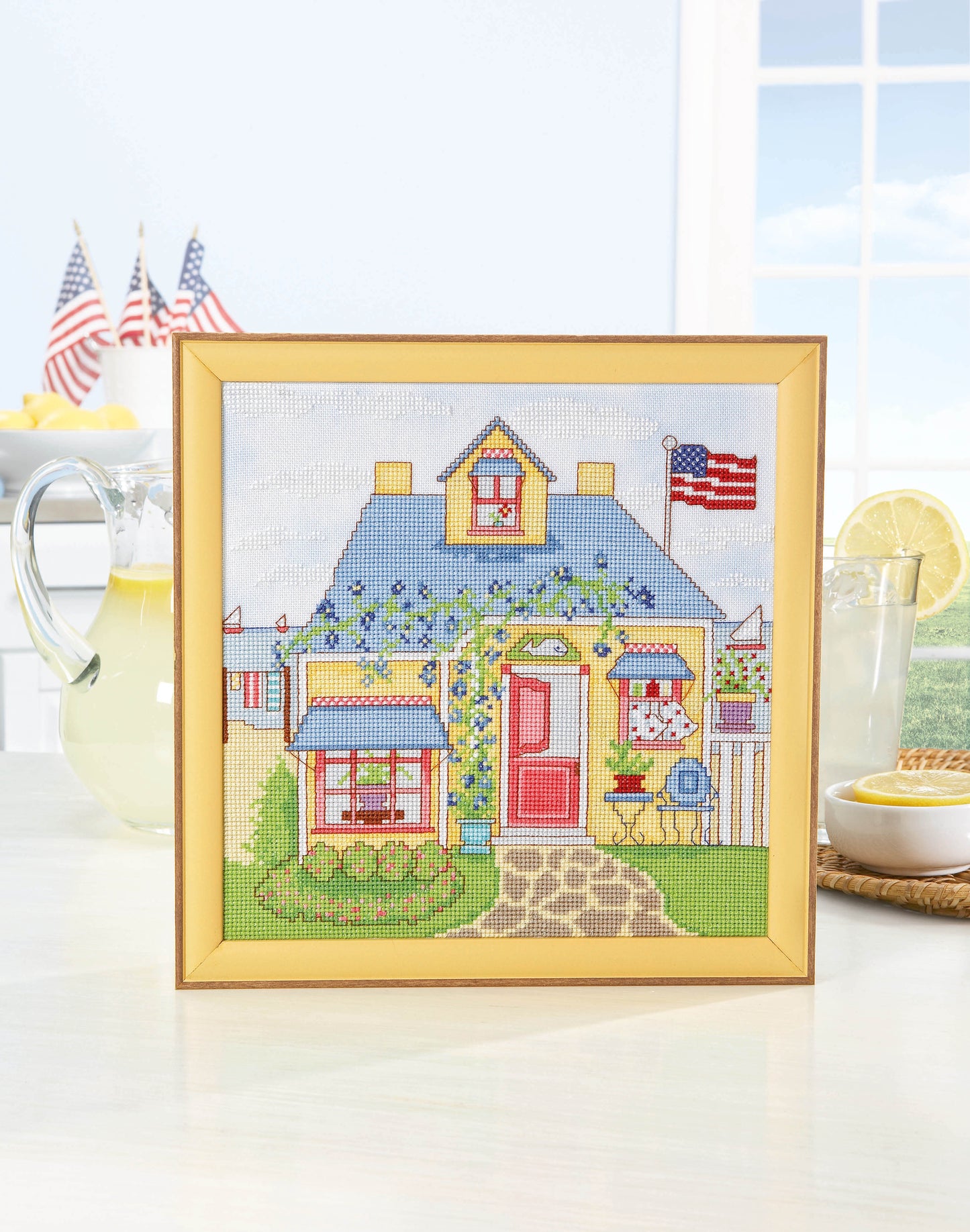 The Leisure Arts Seasonal Cottages Cross Stitch features a yellow house with an American flag, displayed on a table with lemonade, lemons, and mini U.S. flags as sunlight streams through the window for a cheerful look.