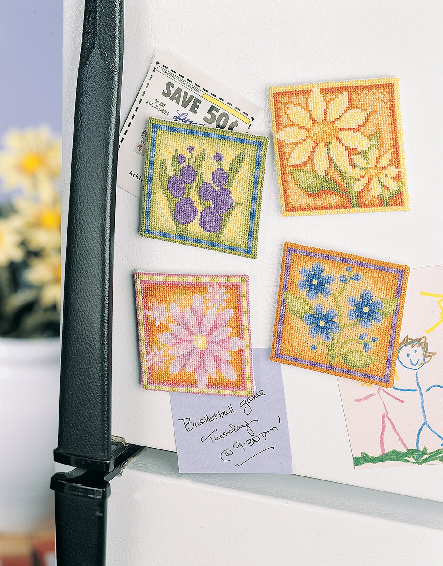 Kitchen Essentials by Leisure Arts—four vibrant cross-stitch flower magnets—hold notes and coupons on a fridge, alongside a child’s drawing and a reminder note. A blurred vase of flowers appears in the background.