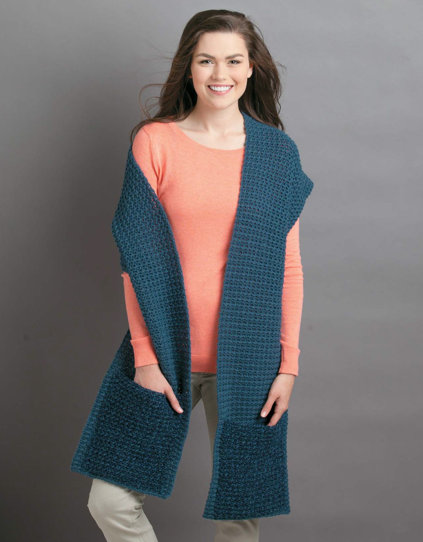 A woman with long brown hair models the Leisure Arts Pocket Shawls Digital Download, featuring a textured dark teal shawl with large pockets over a coral sweater and light pants, standing and smiling against a gray background.