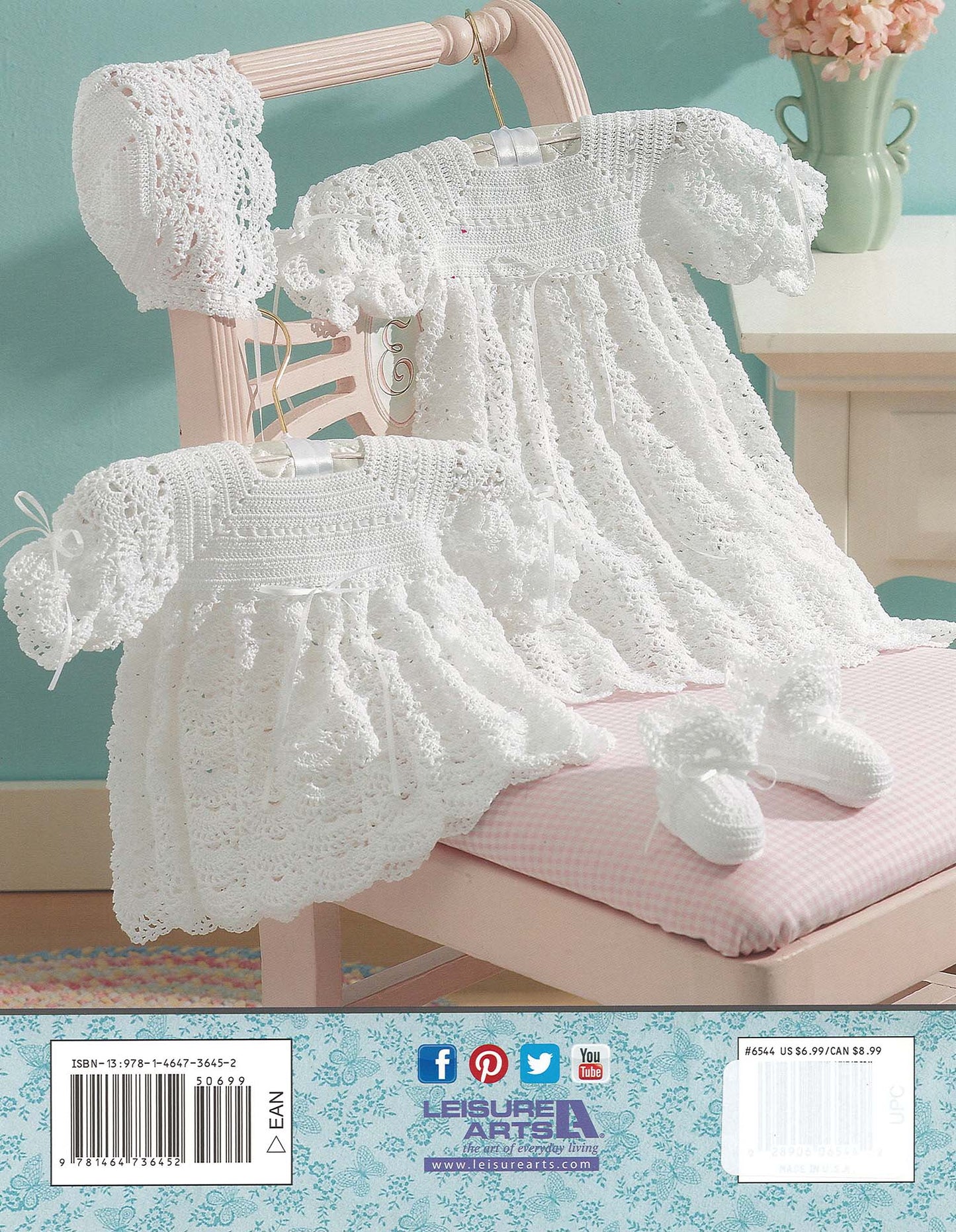 Two white lacy crocheted baby dresses hang on a pink chair, with a matching christening set—bonnet and booties—from Leisure Arts’ Christening Sets: Celebrate Your Newborn With an Heirloom Outfit! All against a teal background.