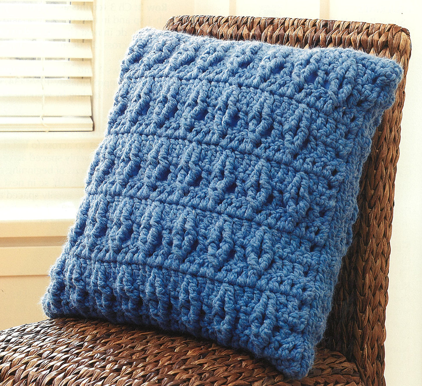 The "Quick Comforts For Home" pillow by Leisure Arts is chunky, blue, and crocheted with super-bulky yarn. It features vertical textures and rests on a woven chair in a bright room with a window and white blinds.
