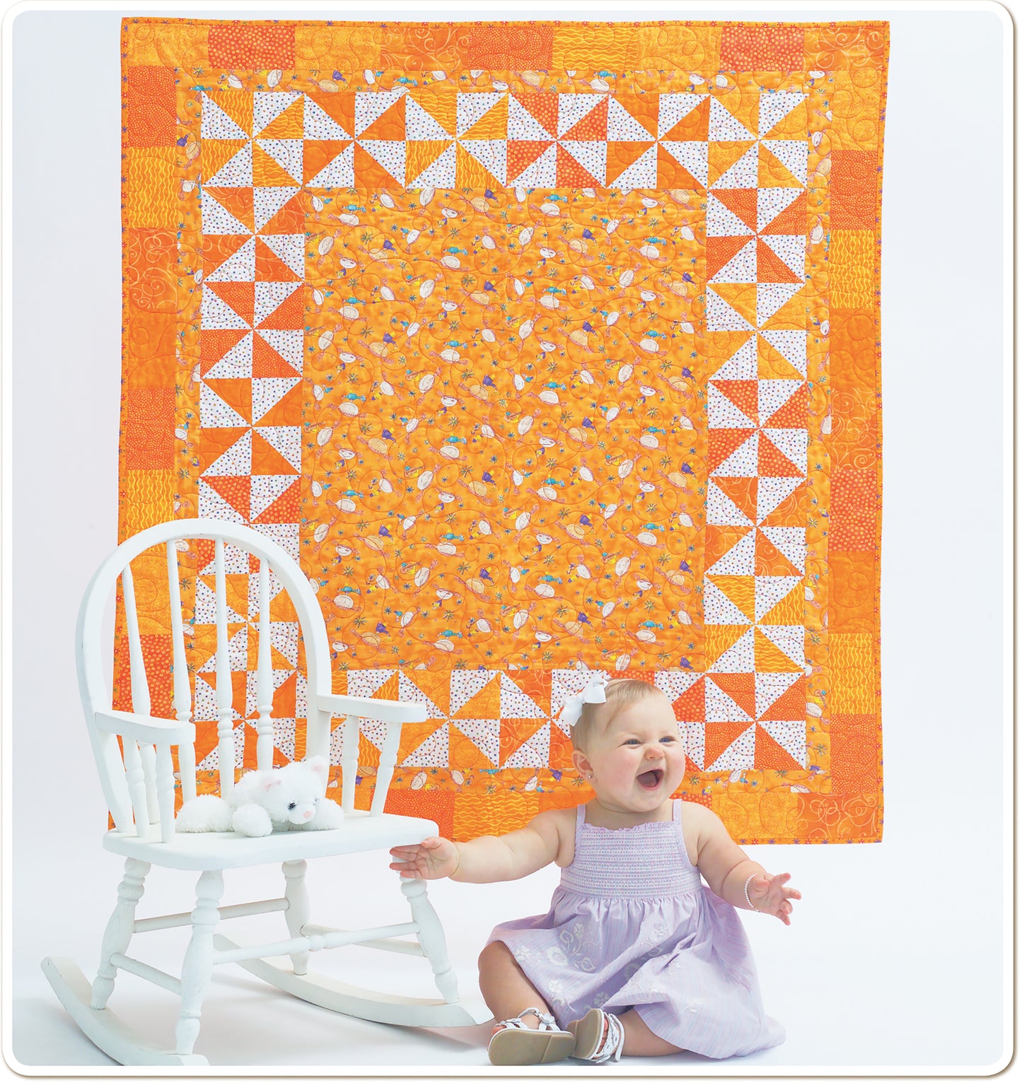 A smiling baby in a purple dress sits by a white rocking chair and stuffed animal, in front of a vibrant quilt from Leisure Arts’ Baby Quilts—Pamper Little Ones with Fast Blankets in Fun Fabrics!, brightening the cozy scene.