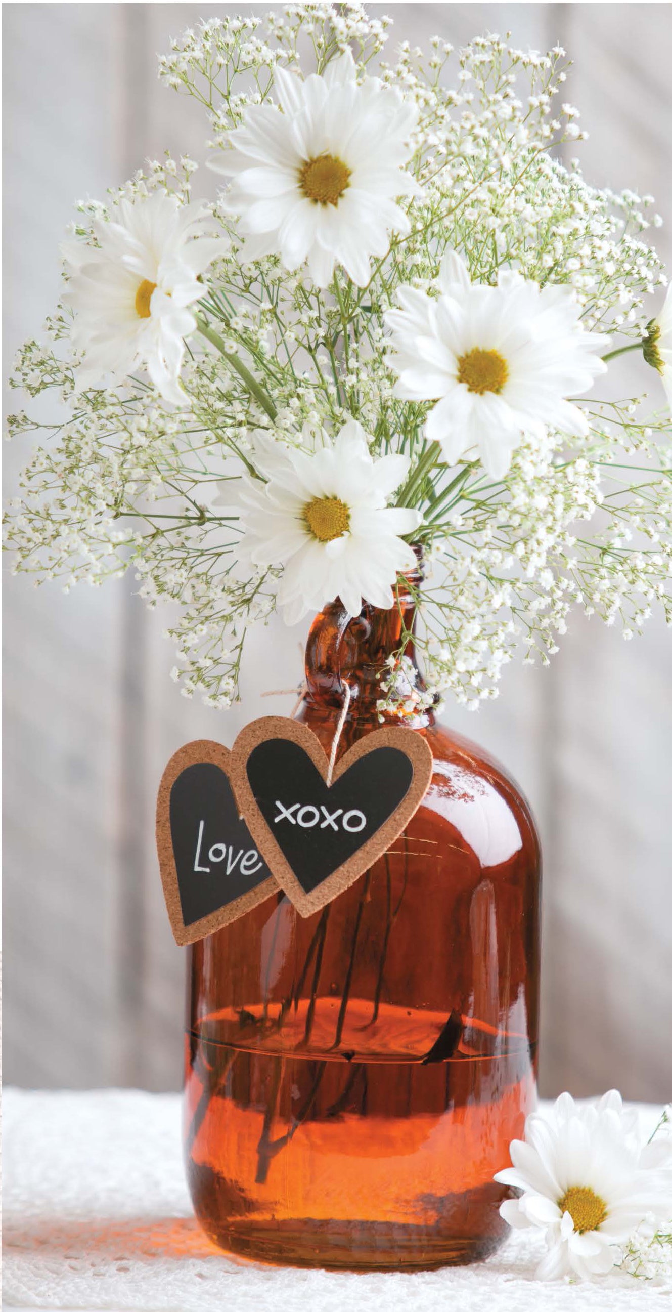 Rustic Wedding Florals by Leisure Arts features a brown glass vase with white daisies, baby's breath, and heart-shaped chalkboard tags—ideal for rustic wedding ideas or DIY decor. A single daisy rests beside the vase.