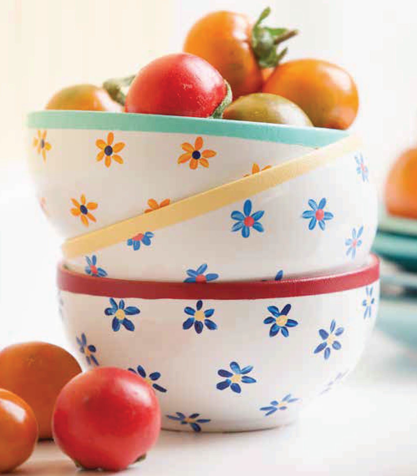 Three white ceramic bowls with blue and orange flower patterns, each filled with cherry tomatoes, are stacked. Extra tomatoes are scattered around like painted rocks from the Painting Basics kit by Leisure Arts for a playful touch.