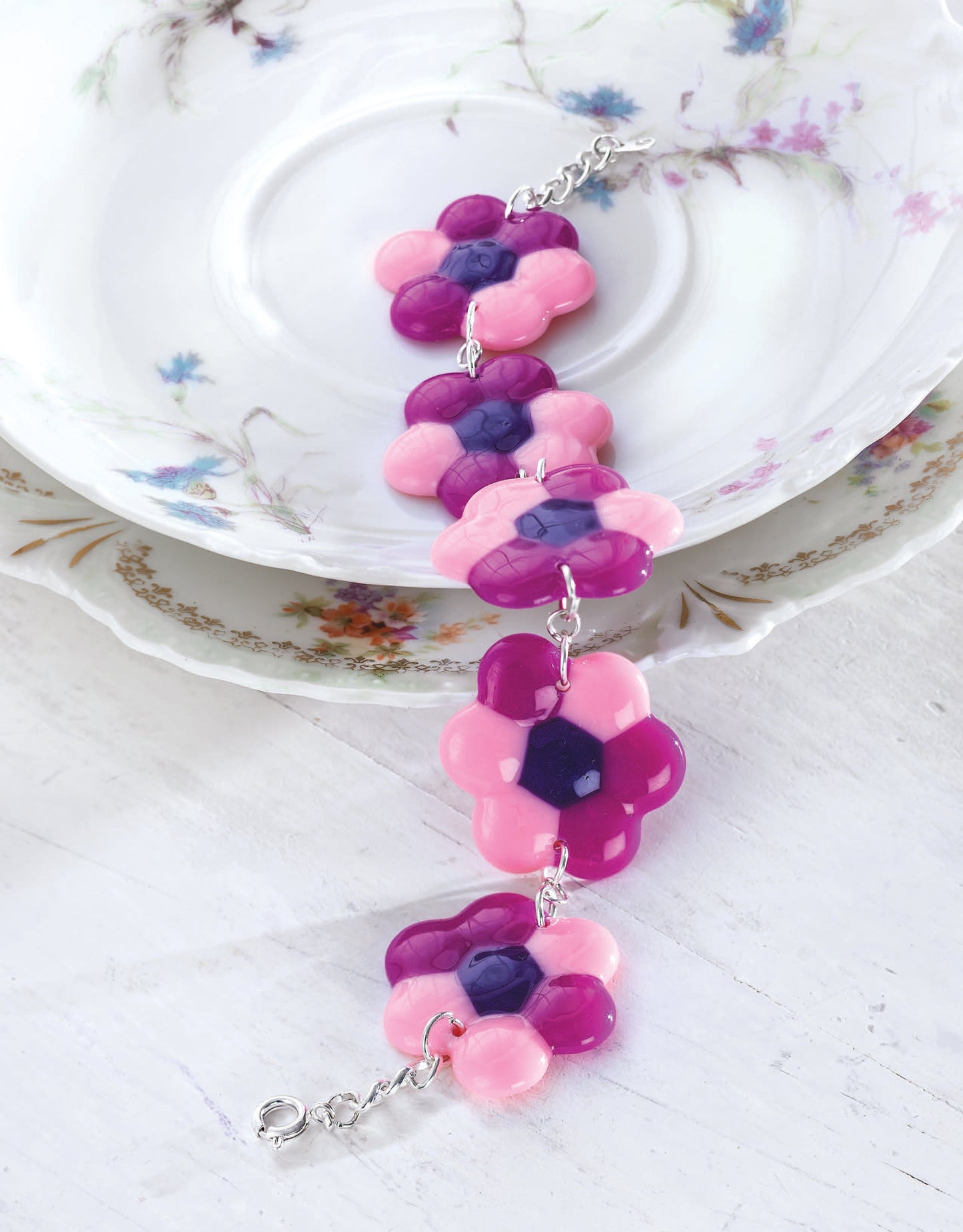 The Leisure Arts Melted Pony Bead Art features shiny pink and purple flower-shaped beads, perfect for beginner crafts or melted bead projects, displayed on floral-patterned plates atop a white wooden surface.