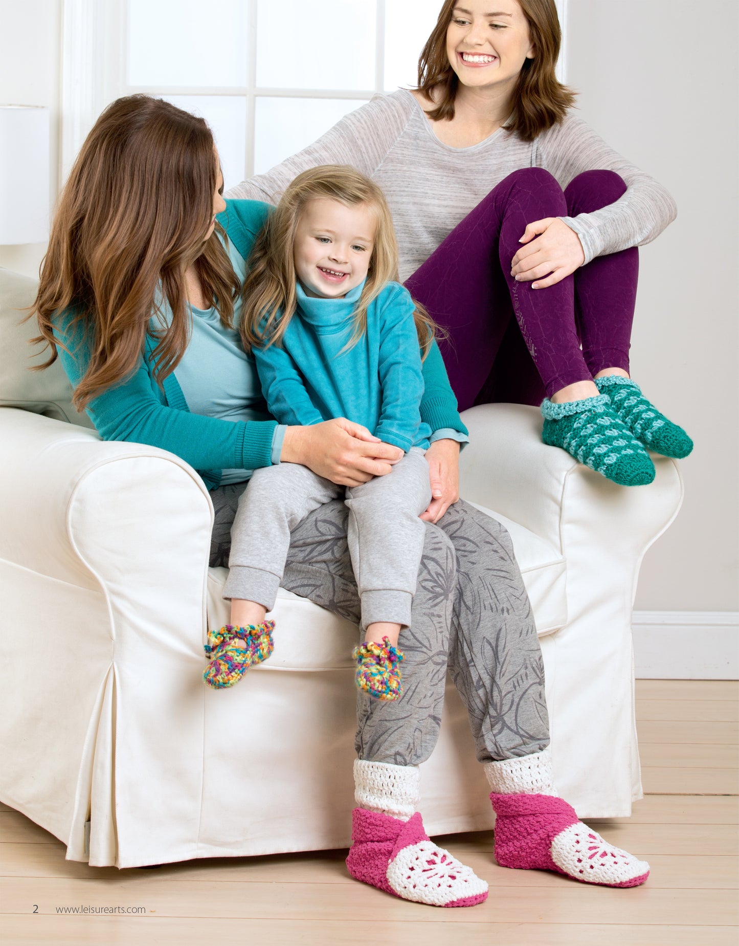 Three people relax on a white couch, smiling and wearing cozy Leisure Arts “Slipper for the Family” crocheted slippers—fun, vibrant house shoes made from a digital pattern download perfect for the whole family.