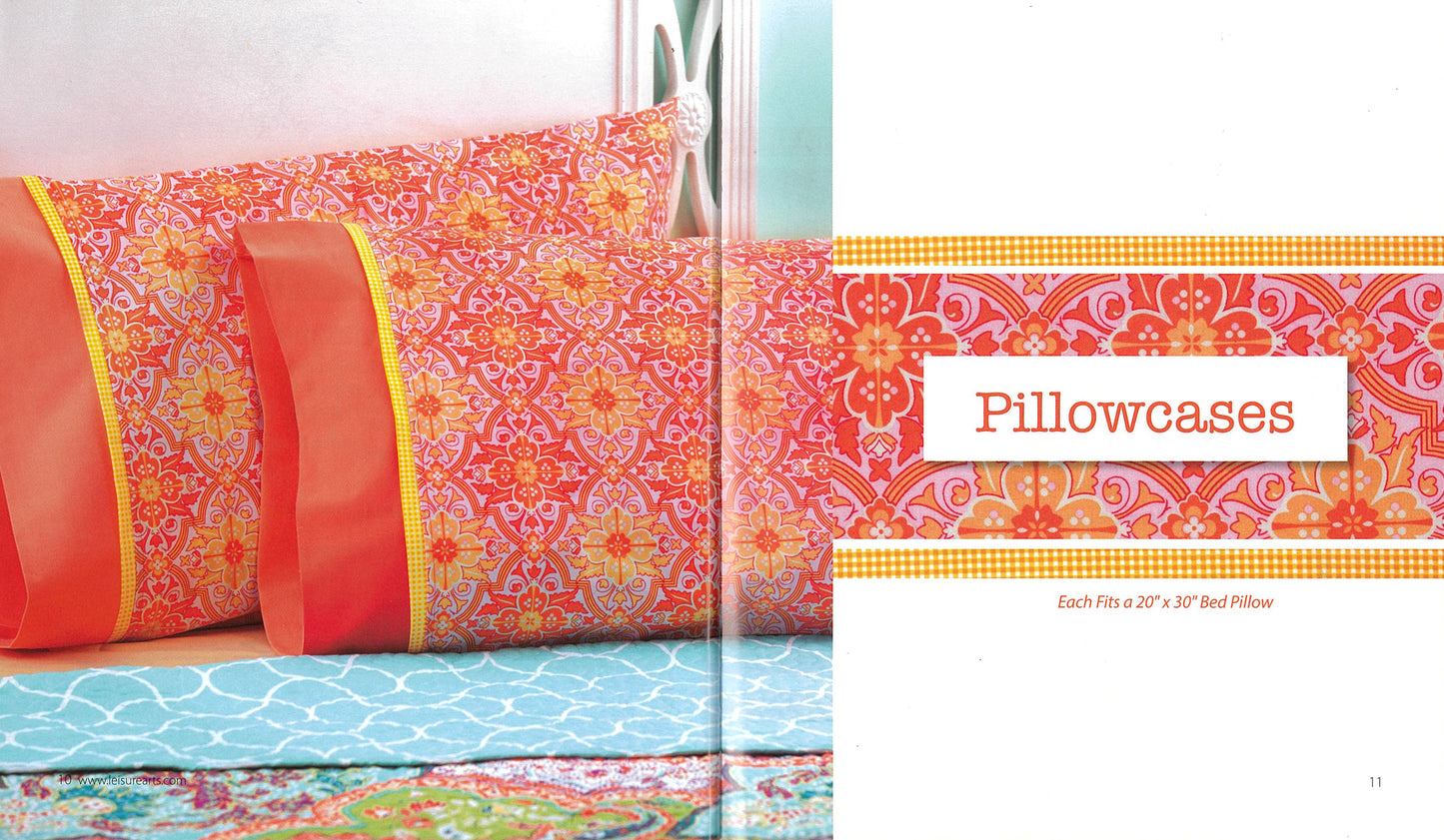 Two colorful Simple Sewing pillowcases by Leisure Arts, featuring orange edges and pink, orange, and yellow patterns, rest on a blue and white quilt. Each fits a 20” x 30” bed pillow.