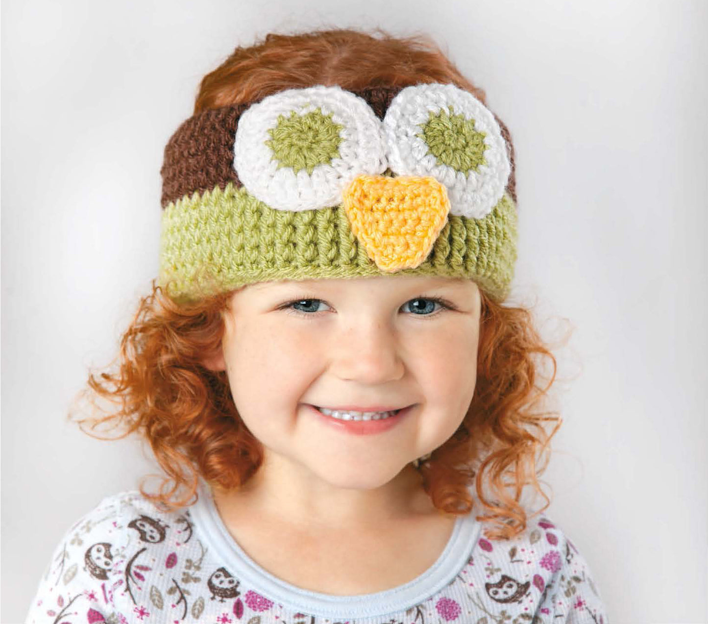 A smiling young child with curly red hair wears the Leisure Arts Little Animals Headgear, an adorable crochet beanie styled as an owl with big eyes and a yellow beak—perfect for fans of cute, easy crochet projects.