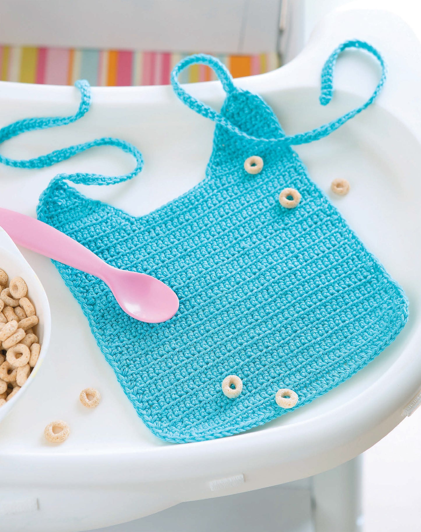 A blue crocheted baby bib from Leisure Arts' "One-Skein Baby Projects" rests on a highchair with cereal and a pink spoon. Colorful stripes in the background make it an ideal handmade gift for newborns.