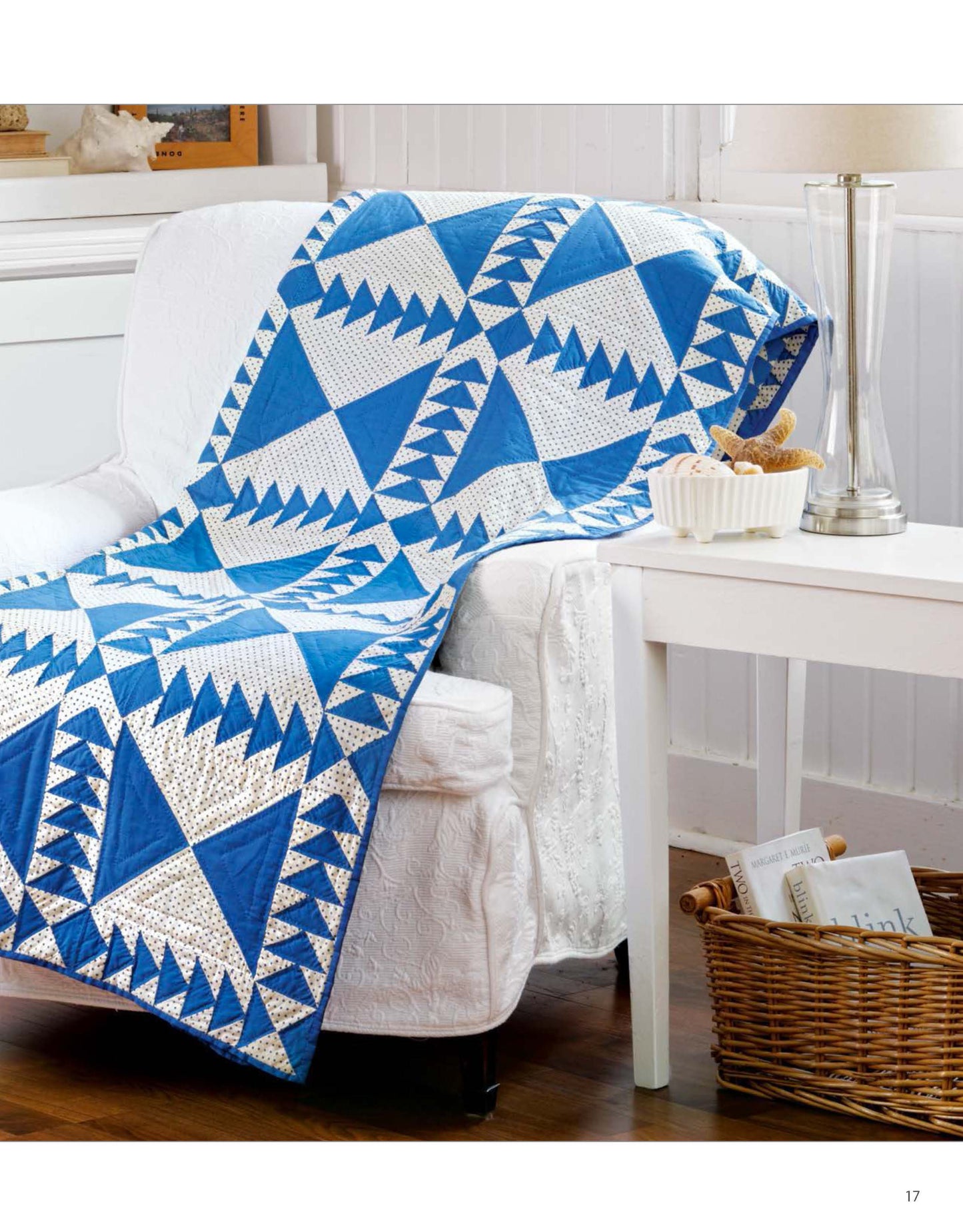 A Classic Quilts blue and white geometric quilt by Leisure Arts is draped over a white couch beside a side table with a tall glass lamp, white dish, and a basket of books and magazines underneath.