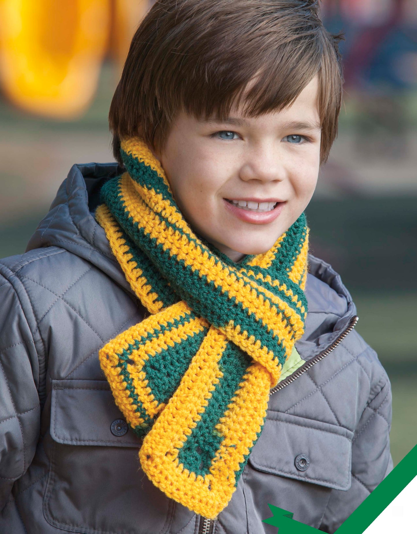 A young boy with brown hair wears a gray quilted jacket and a crocheted green and yellow striped scarf made from Leisure Arts' Home Team Gear, smiling outdoors.