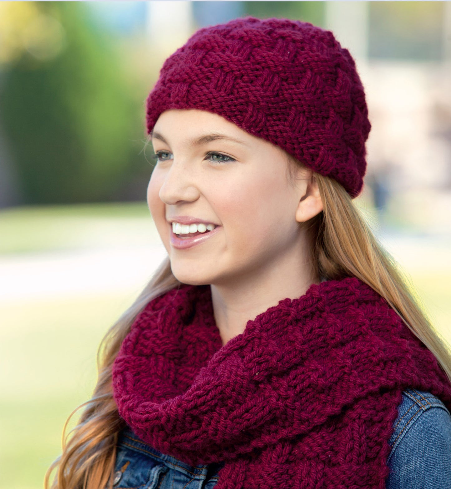 A smiling young person in a matching knit burgundy hat and scarf, perfect for “Hats and Scarves for Kids Digital Download” by Leisure Arts, stands outdoors with long hair and a denim jacket against a blurred green background.