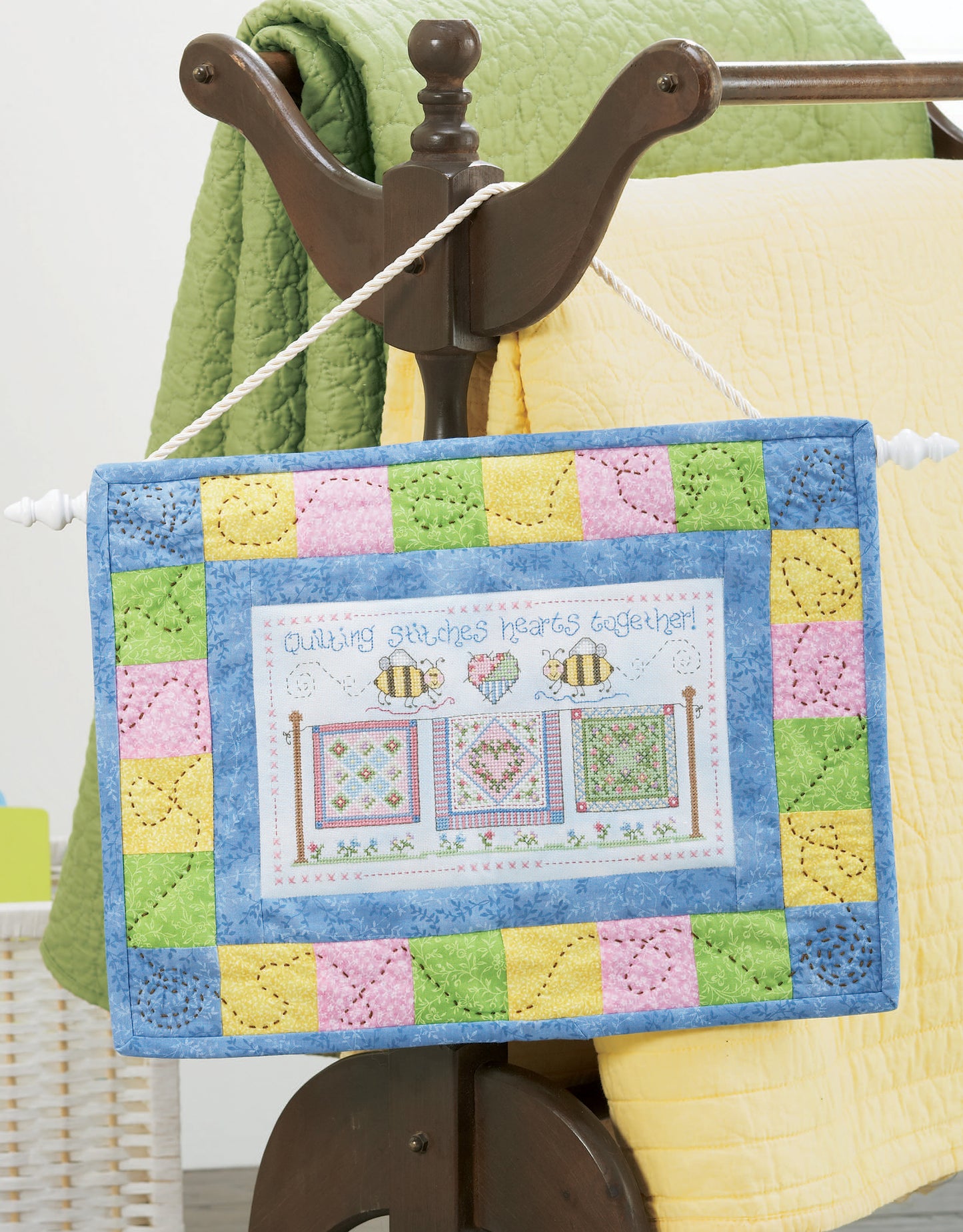 The Loving Hearts Cross Stitch by Leisure Arts features bees, small quilts, hearts, and “Quilting stitches hearts together!” on a colorful quilted sign with patchwork borders—a charming home decor piece. Yellow quilt and green blanket in the background.