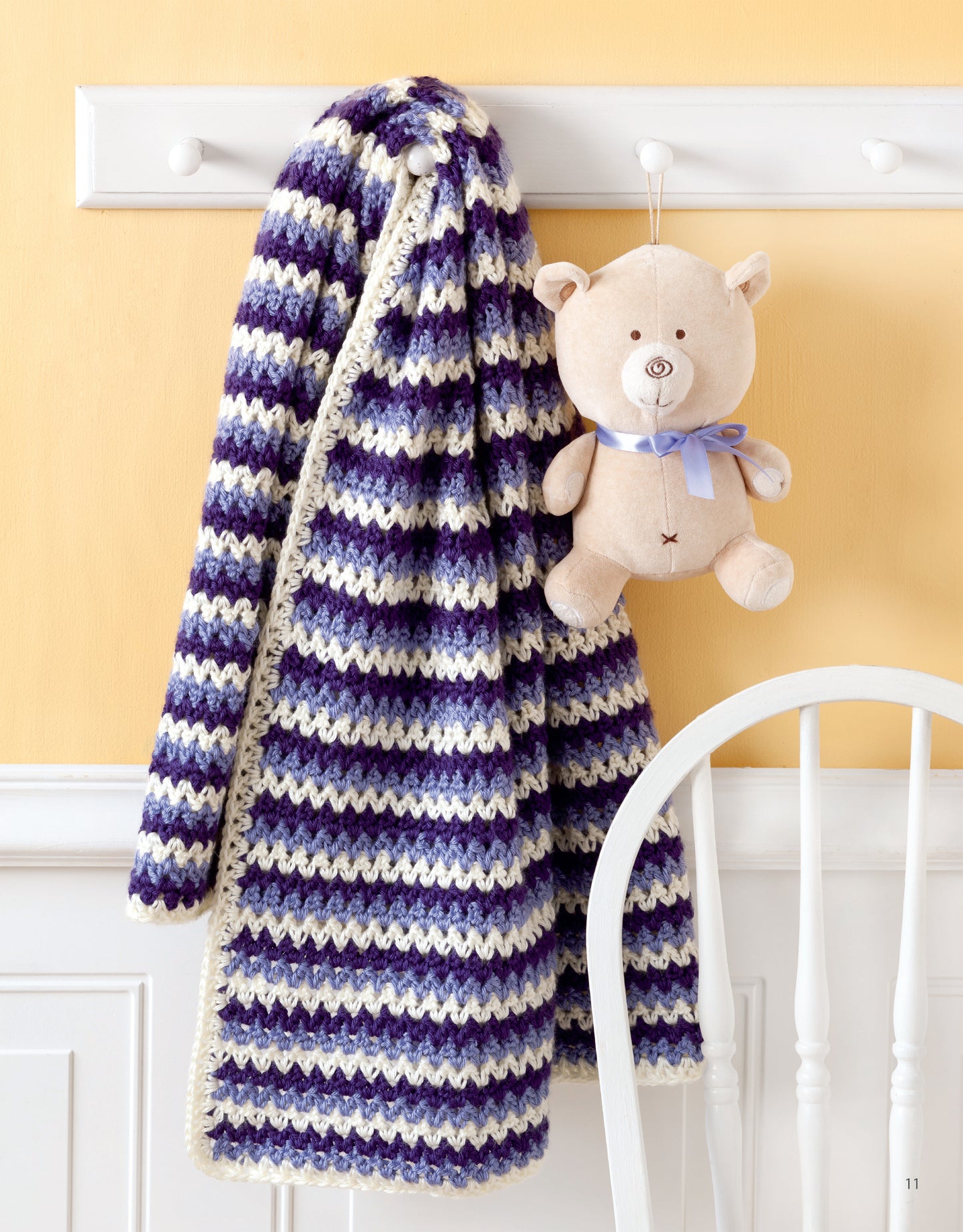 A striped purple and white afghan from Leisure Arts' "Sweet Stripes for Baby—Make 3 Color Afghans" hangs with a beige teddy bear with a blue ribbon on wall hooks above a white chair, set against a yellow wall.