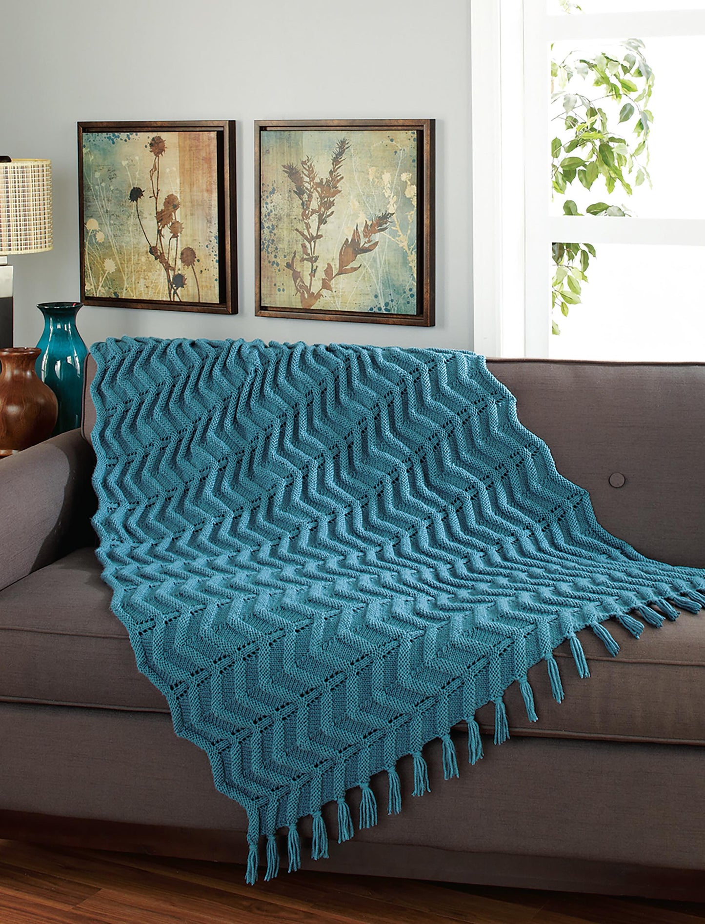 A Knit Classic Afghans throw by Leisure Arts in blue chevron pattern drapes over a gray couch in a cozy living room with framed botanical art, a green vase, and natural light streaming through the window.