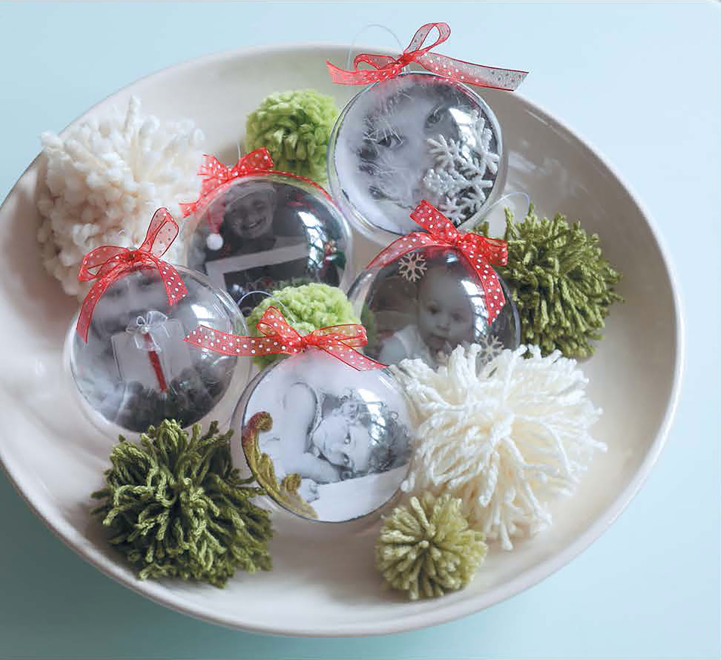 A white bowl holds Leisure Arts’ Open House Merriment clear photo ornaments, each adorned with red bows and green and white yarn pom-poms and featuring black-and-white photos inside—perfect festive accents to brighten up your holiday décor.