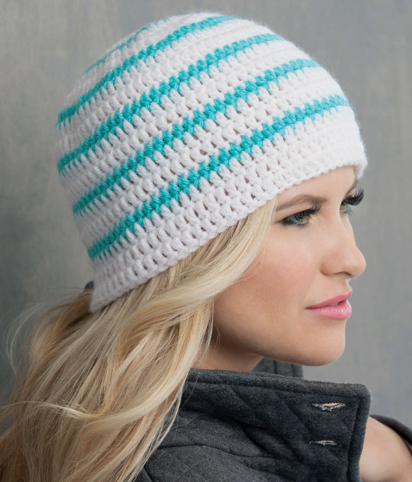 A woman with long blonde hair wears a crocheted white beanie with turquoise stripes, made from beginner patterns in "Learn to Crochet the Easy Way" by Leisure Arts. She is in profile, facing right, wearing a dark gray jacket against a neutral background.