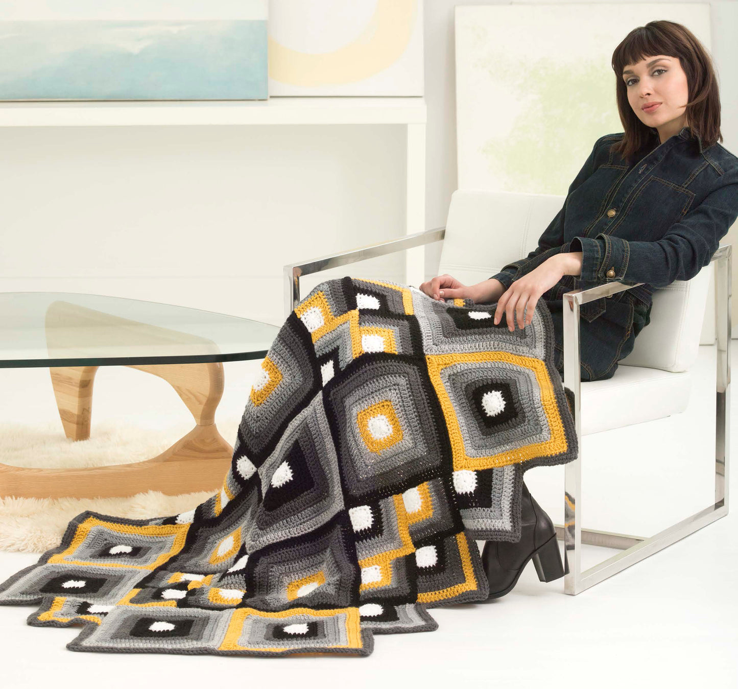 A woman relaxes in a modern chair by a glass table, wrapped in the Mindful Mandala Afghans from Leisure Arts, featuring black, gray, white, and yellow geometric squares inspired by mandala crochet patterns.