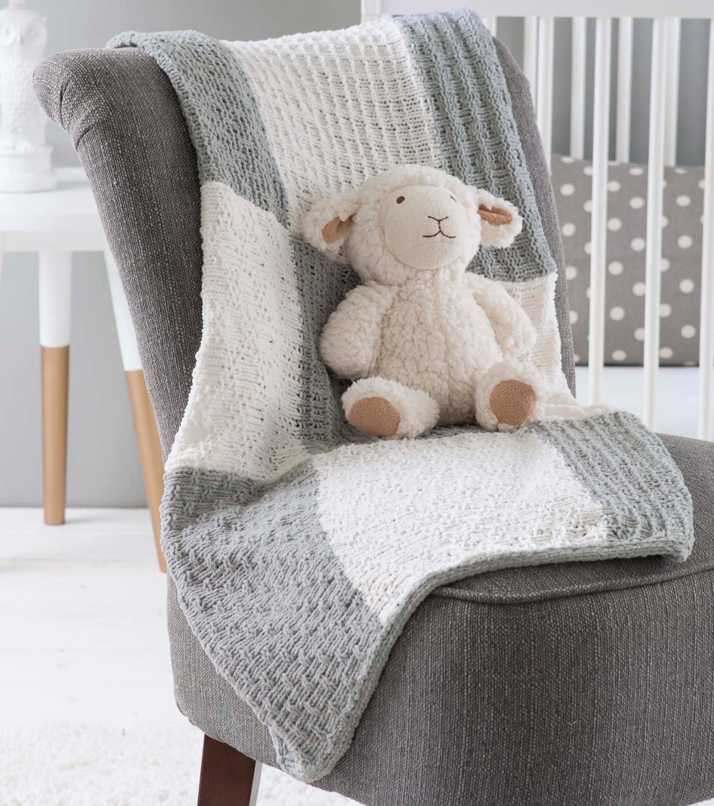 A plush lamb toy sits on a gray chair draped with the Loom Knit Samplers afghan by Leisure Arts, featuring various stitch patterns; a white crib with polka dot bedding can be seen in the background.
