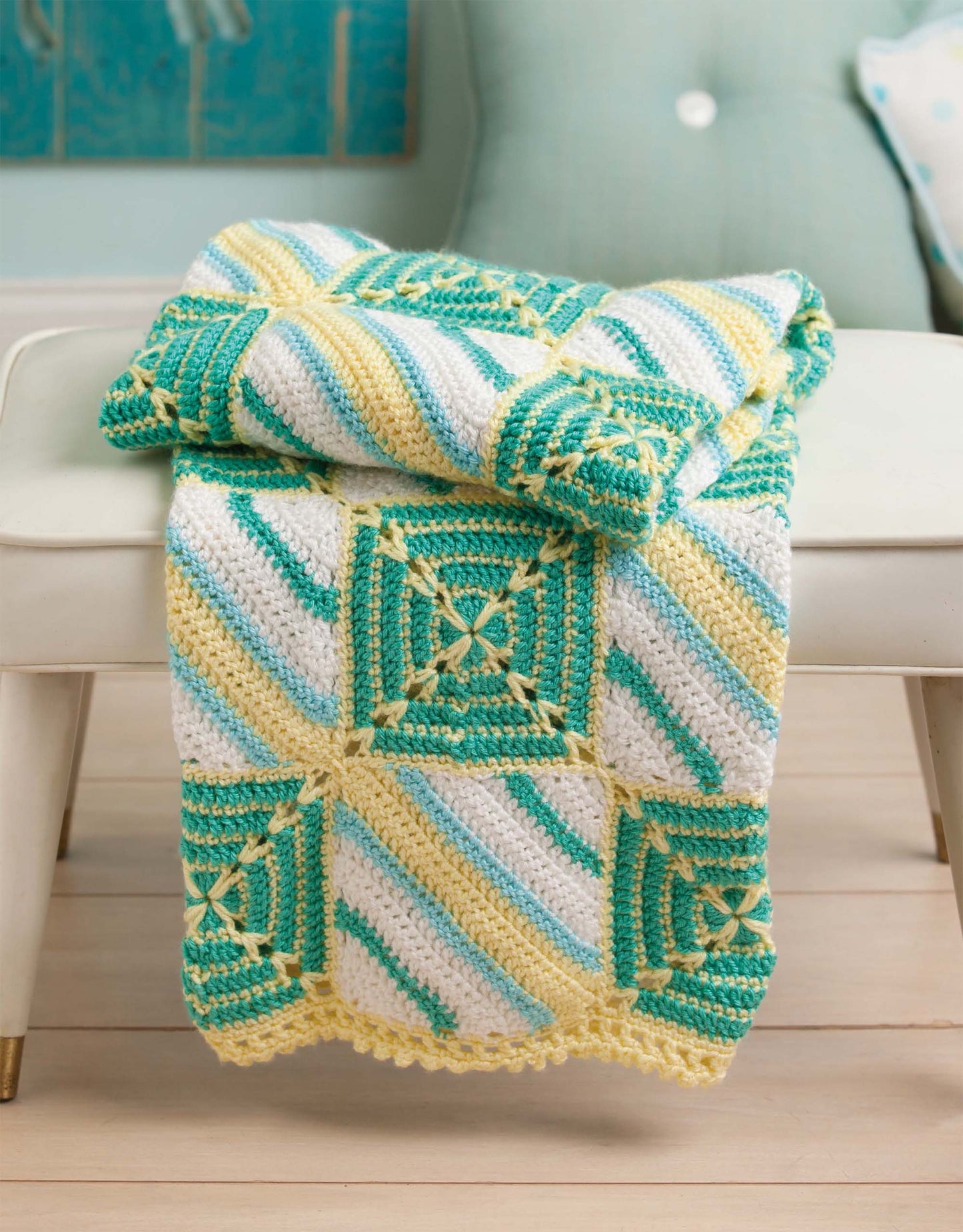 The Mix & Match Baby Sampler crocheted blanket by Leisure Arts, featuring green, yellow, and white granny squares, is draped over a white bench in a bright, cozy room.