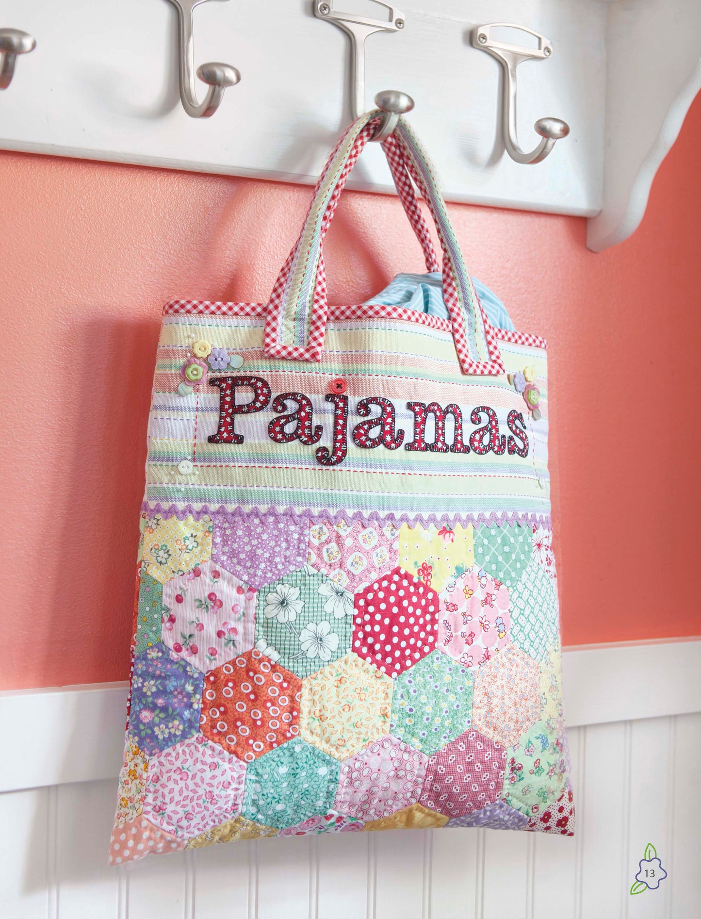 A colorful patchwork bag from the Salt Water Taffy Collection by Leisure Arts hangs from a hook on a white shelf against a pink wall. Reminiscent of heirloom quilts, the bag features pastel and floral hexagon patterns crafted using English paper piecing techniques.