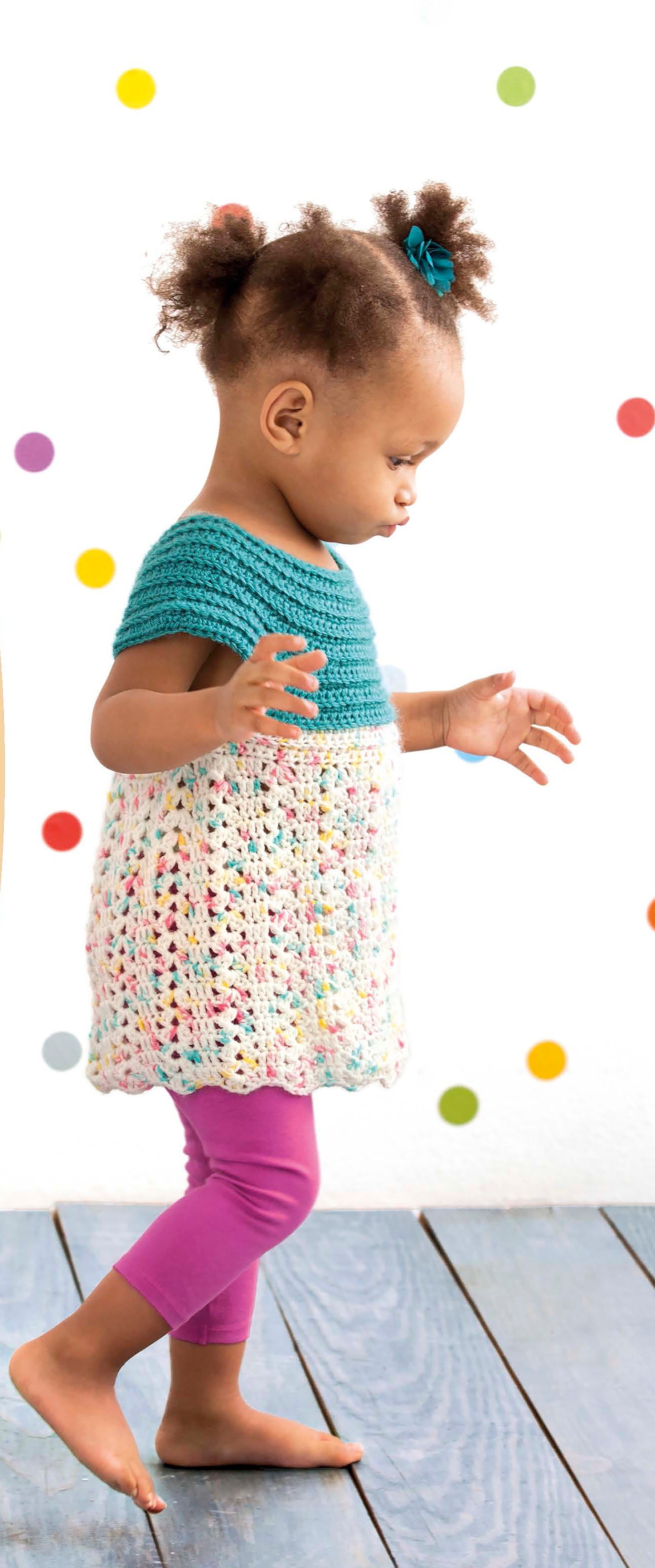 A young child in a Lacy Little Girl Dress by Leisure Arts and purple leggings walks barefoot on a wooden floor, with colorful polka dots on a white wall in the background.