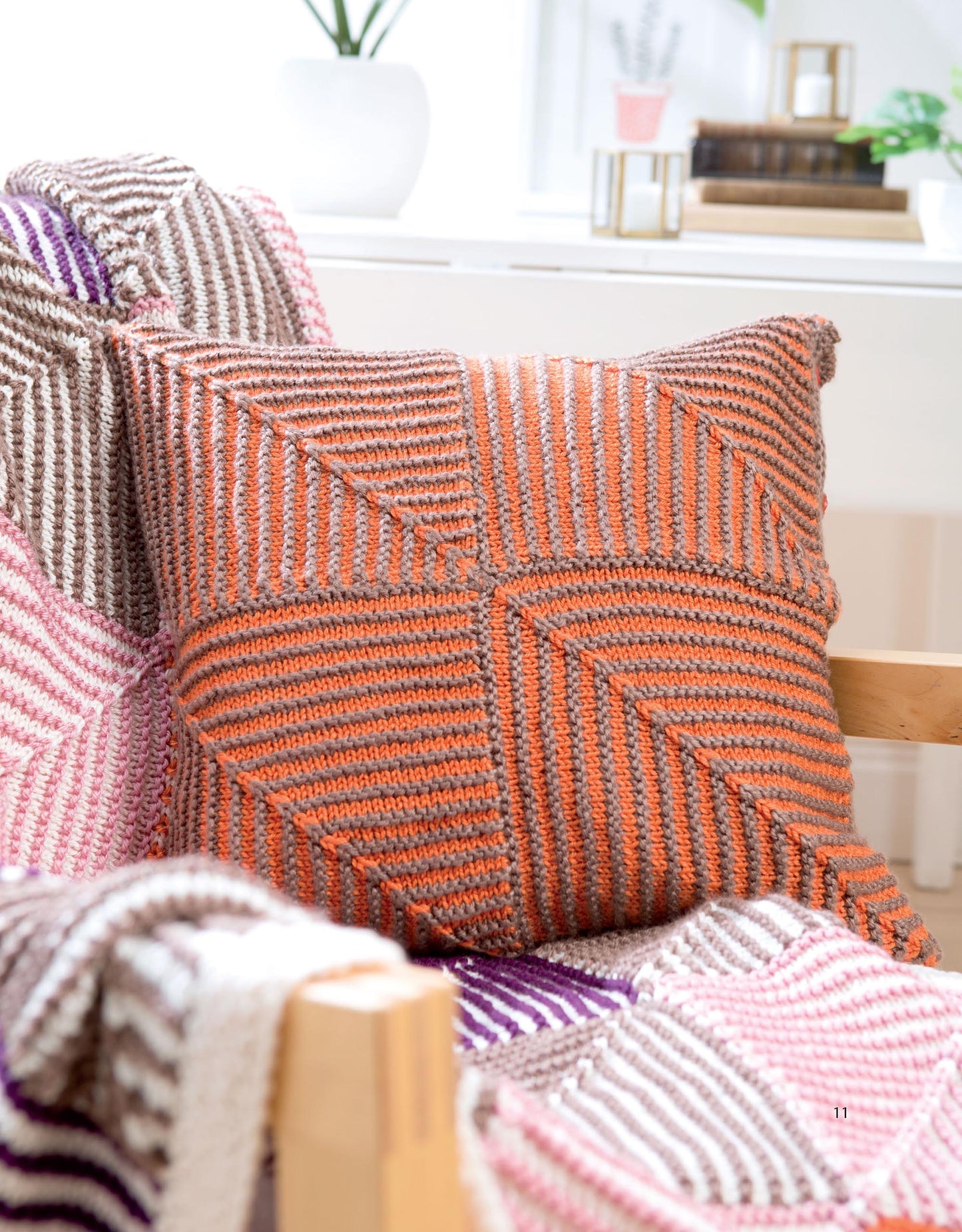 A cozy room features a chair with a colorful patchwork afghan topped by a pillow knit in orange and brown stripes, inspired by the geometric look of mitered squares from the "Mitered Square Knitting Digital Download" by Leisure Arts.