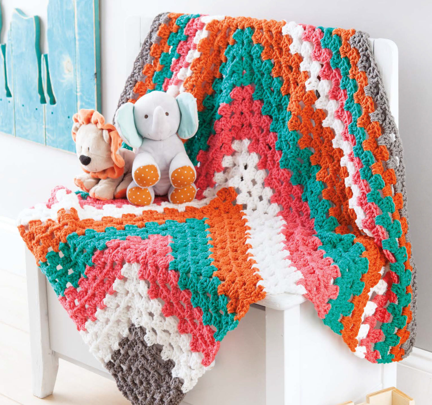 The Leisure Arts Color-Block Baby Blankets, crocheted with medium weight yarn in white, coral, pink, teal, and gray chevron stripes, drape playfully over a white chair and complement stuffed lion and elephant decor.