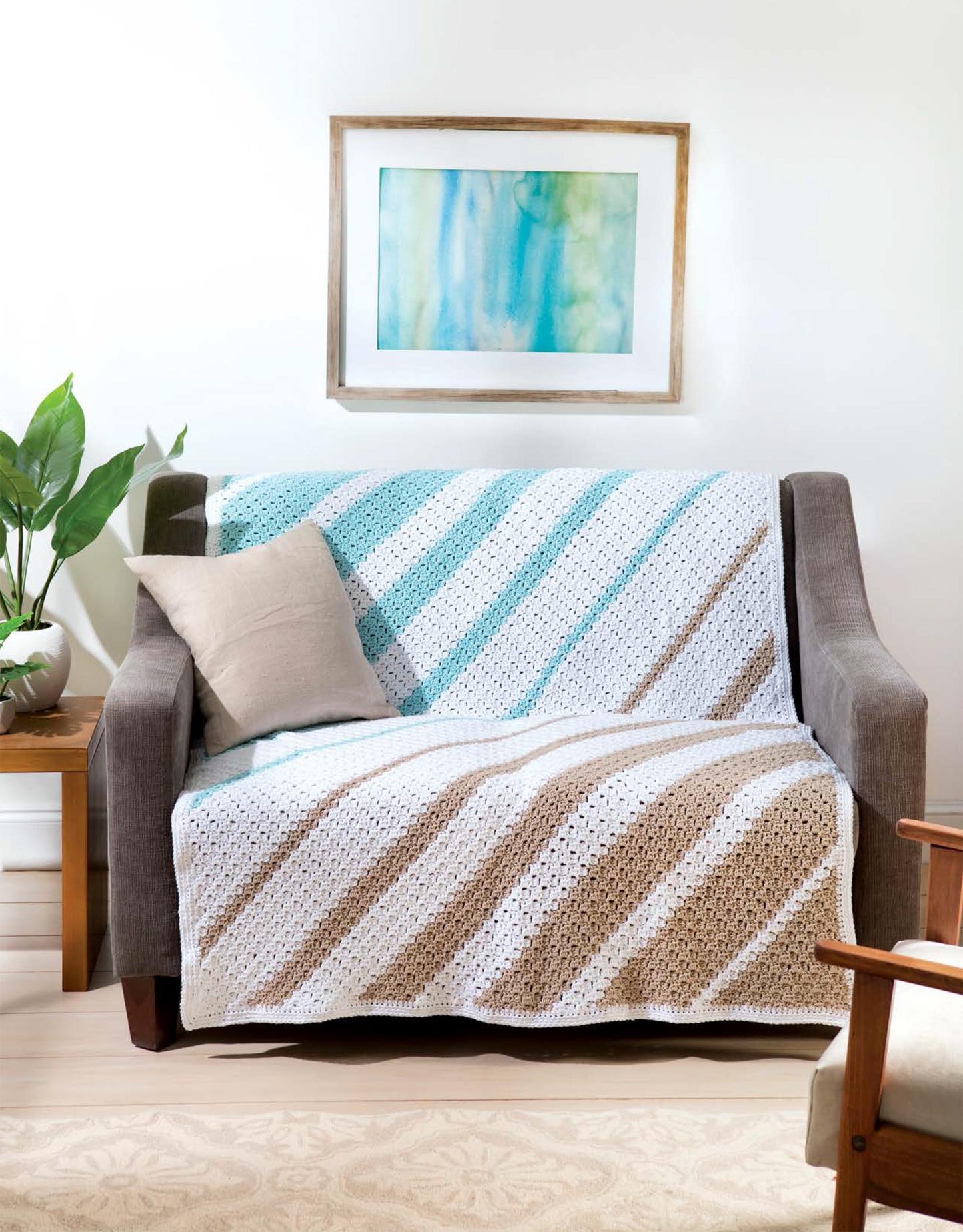 A gray loveseat is draped with a Leisure Arts Cotton Blanket & Throw, crafted from medium weight yarn in white, teal, and tan diagonal stripes. A beige pillow sits on one side; a plant, table, and abstract art accent the cozy living space.