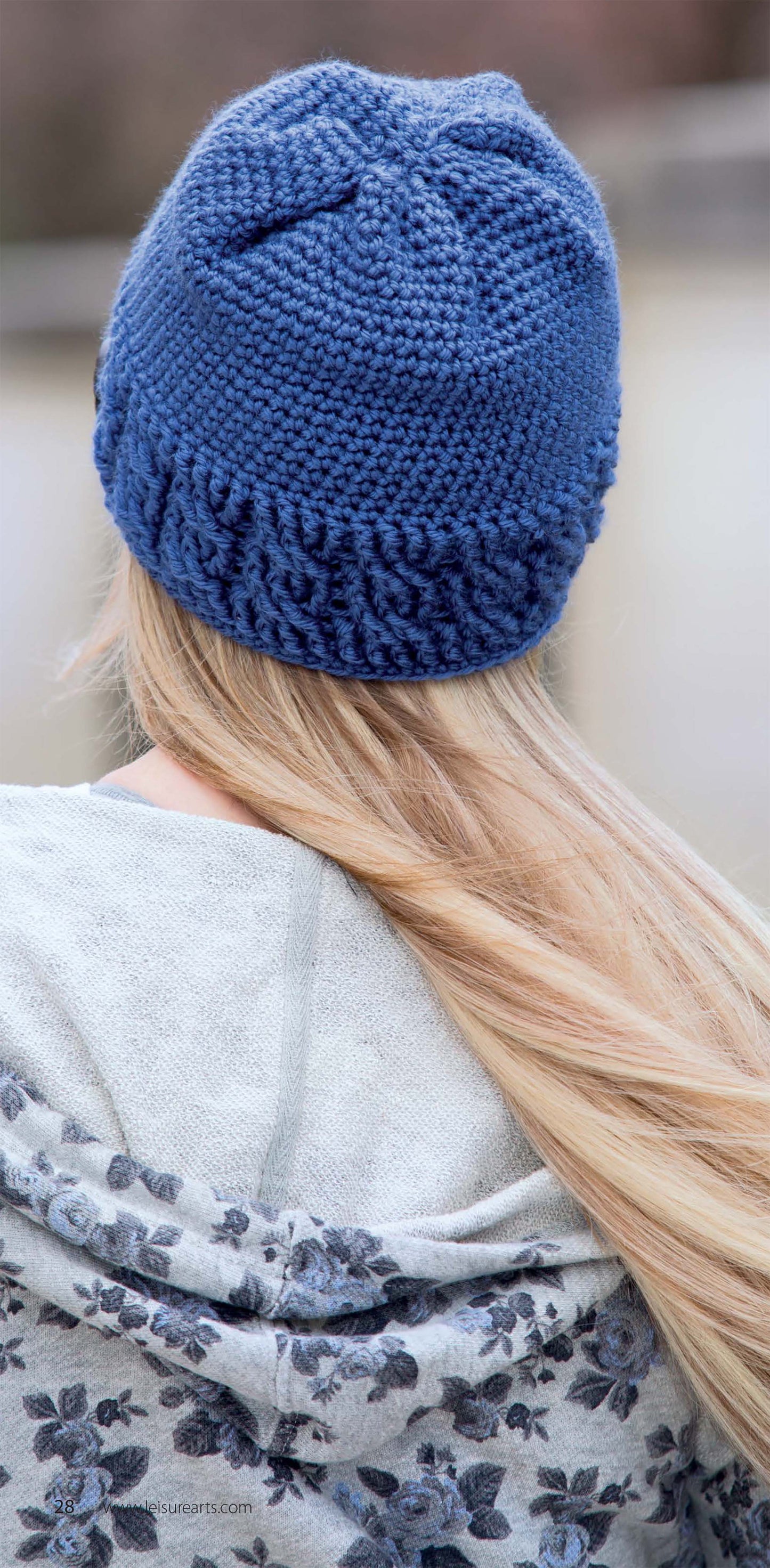 A person with long blonde hair is seen from behind, wearing a blue Messy Bun Hat by Leisure Arts and a grey patterned jacket. The softly blurred background highlights the stylish hat.