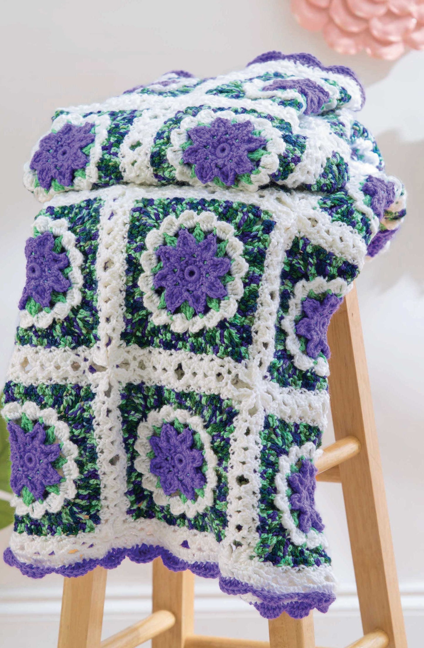 The Grannies In Bloom Digital Download by Leisure Arts features a crocheted blanket with purple, green, and white floral granny squares, scalloped edges, and detailed stitches displayed on a wooden stool. Medium weight yarn is recommended.
