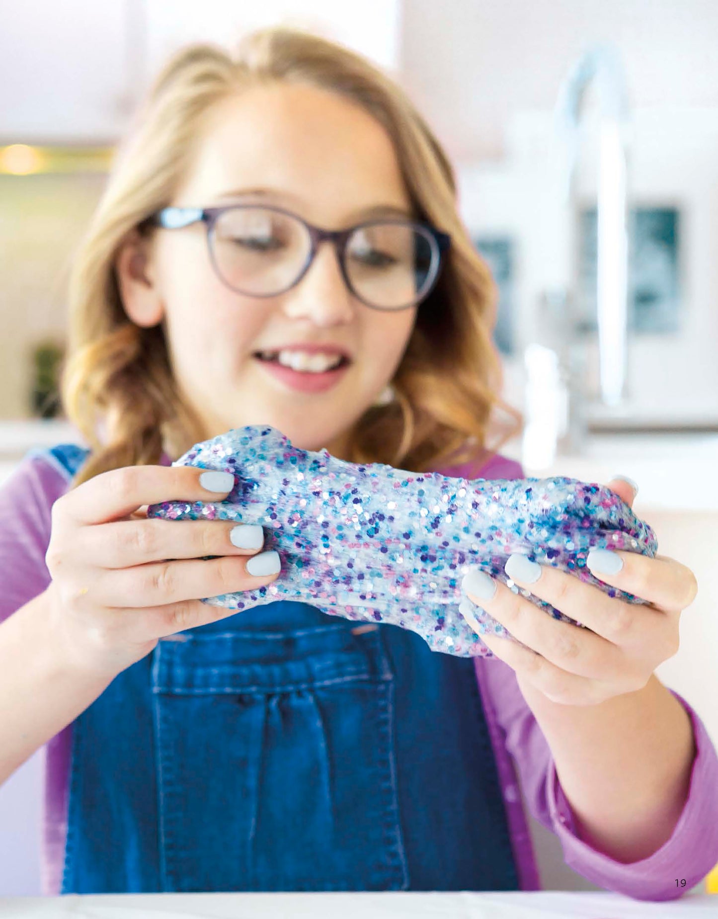 A young girl with glasses and light brown hair, wearing a blue apron, smiles while playing with sparkly purple and blue slime—perfect fun inspired by Leisure Arts' "Slimed DIY—A guide to making Slime at home!" digital download.
