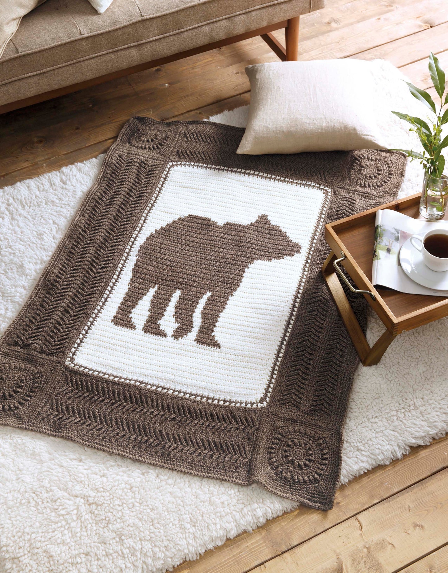 The Wildlife Cable Afghans by Leisure Arts features a crocheted blanket with a brown bear silhouette, shown on a white rug beside coffee and books—ideal for animal-themed decor lovers seeking cozy throw blankets.
