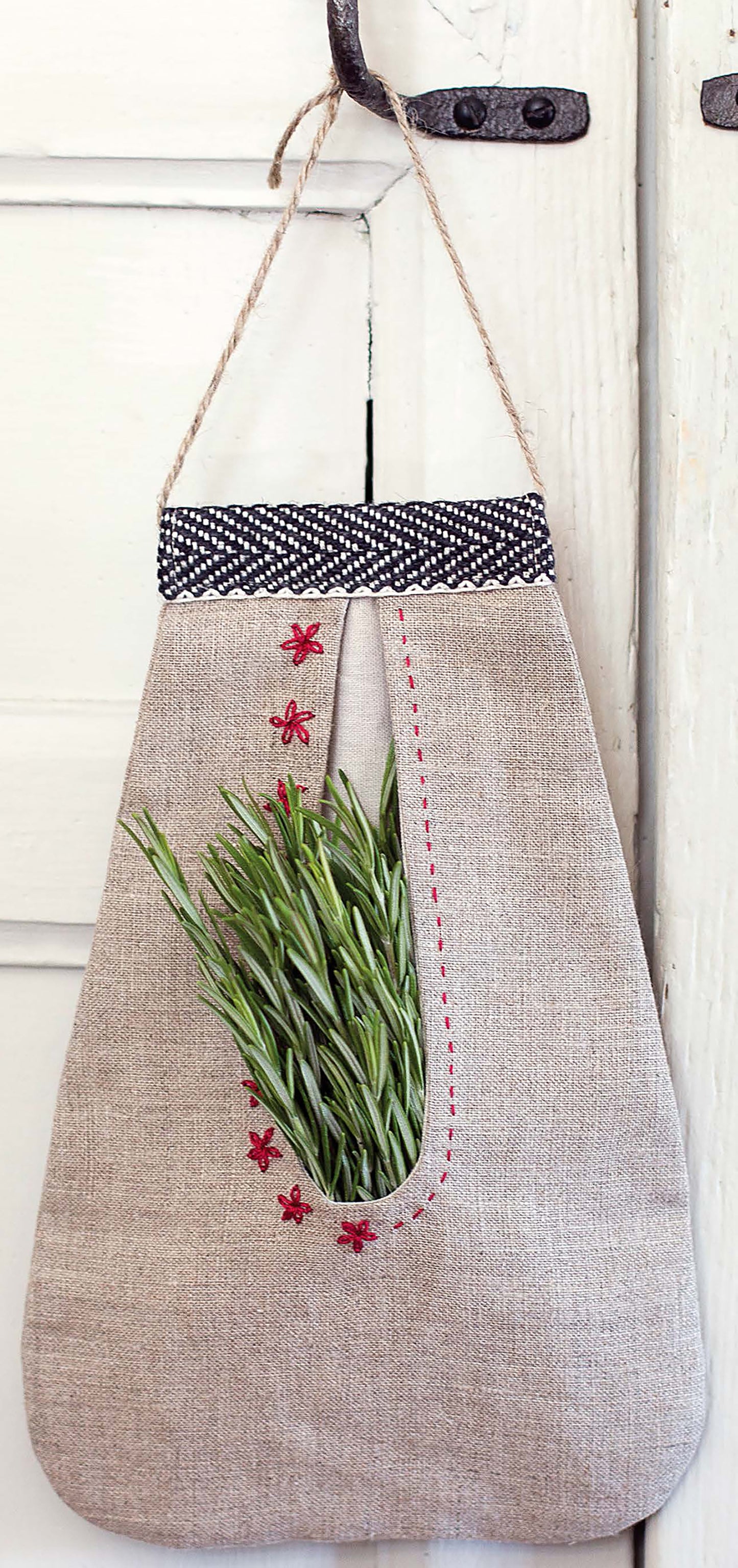 A linen hanging bag from Leisure Arts’ “Sewing Home—11 Useful & Beautiful Decor Projects,” with floral trim and sashiko embroidery, holds rosemary sprigs and hangs on a white wooden door.