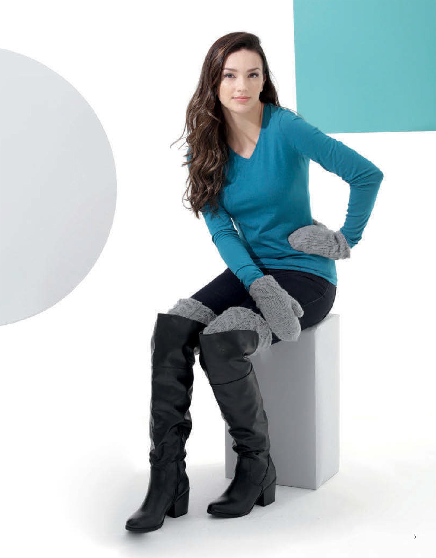 A woman with long brown hair, wearing a teal long-sleeve shirt and Leisure Arts Knit Accessories—gray mittens and leg warmers—sits on a white block amid geometric shapes in a minimalist white setting.