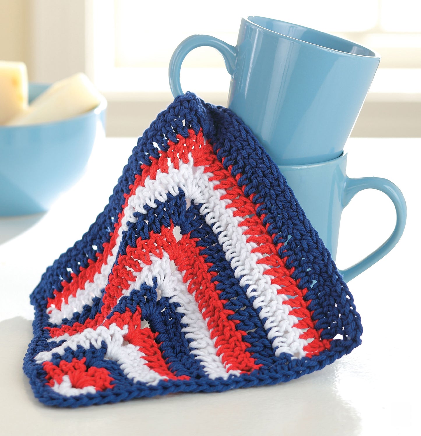 Two stacked blue mugs draped with a vibrant red, white, and blue crochet cloth from Leisure Arts' "Quick & Colorful Dishcloths Designs to Brighten Any Kitchen" digital download. A blue bowl with light items is in the background on a white surface.