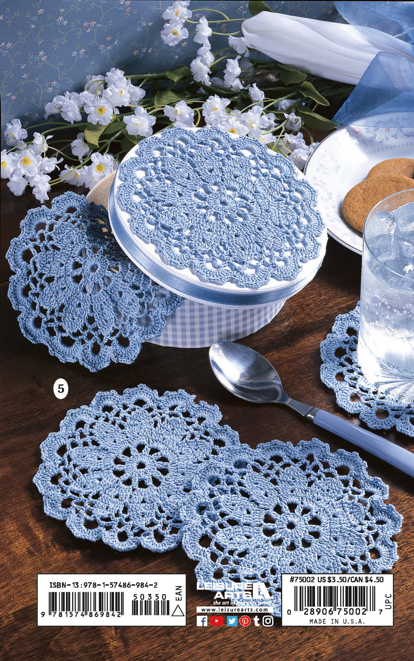 Four light blue crocheted doilies made from "Thread Crochet—A Beginner's Guide to 9 Projects" by Leisure Arts are displayed on a table beside cookies, a drink, and flowers, perfect for beginners or as decorative heirloom accents.