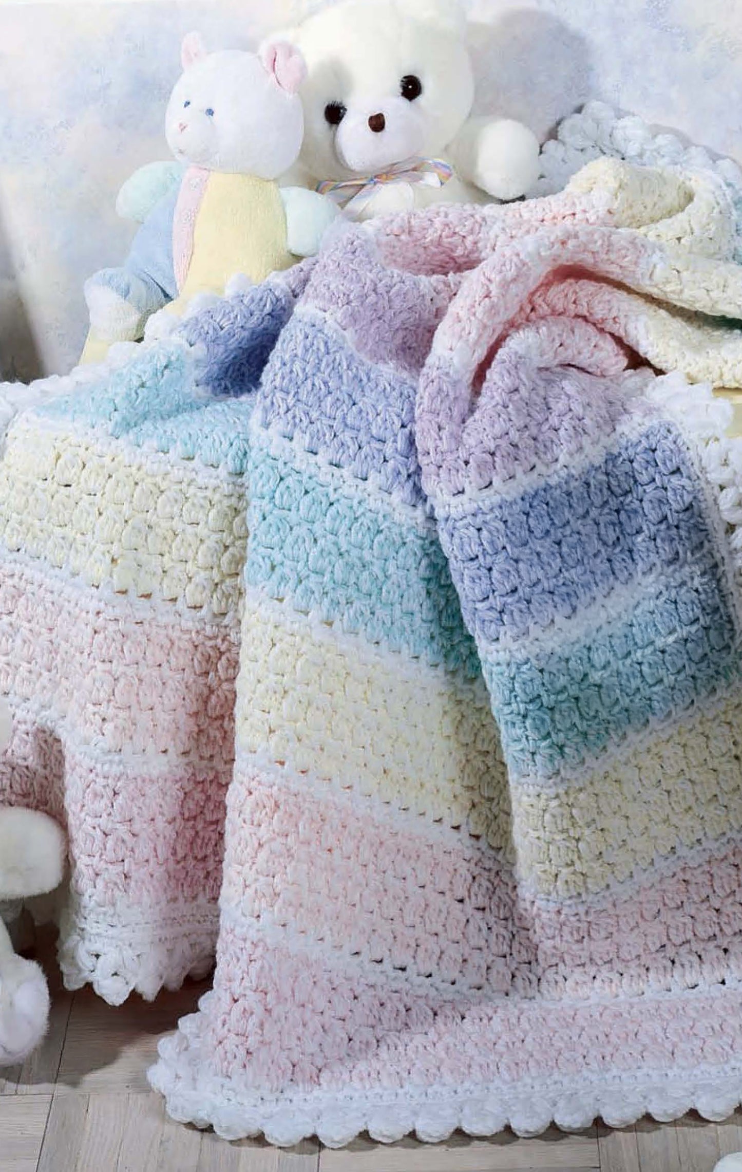 The Snuggletime Baby Afghans by Leisure Arts, featuring pastel stripes of pink, blue, purple, green, and white, is draped on a chair with plush teddy bears and a bunny toy—ideal for crochet lovers.