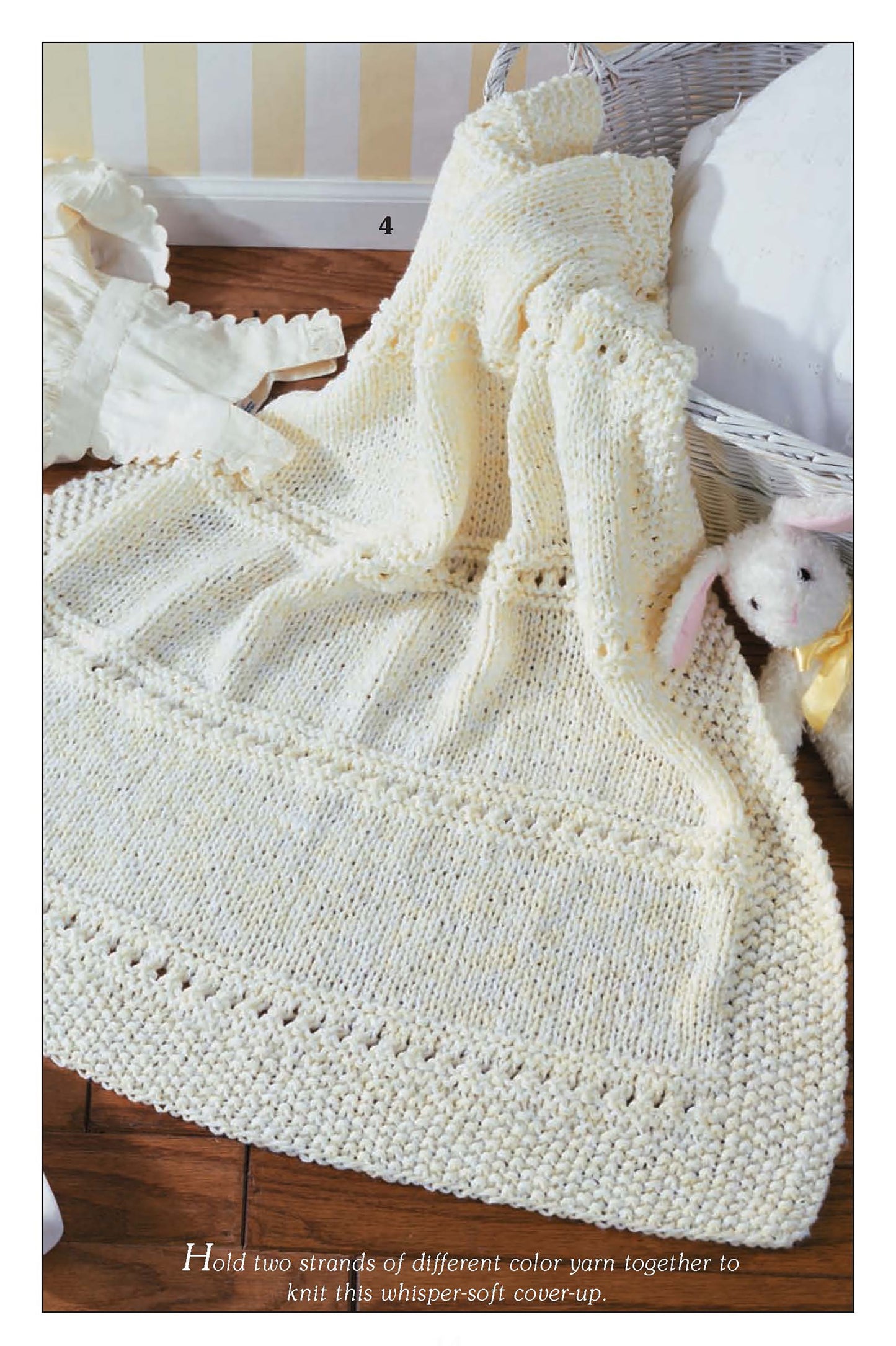 The "So Sweet Baby Afghans" by Leisure Arts, a cream-colored knitted blanket ideal for beginner projects, is draped over a basket next to a plush white lamb on a wooden floor, with soft light and pale striped walls in the background.