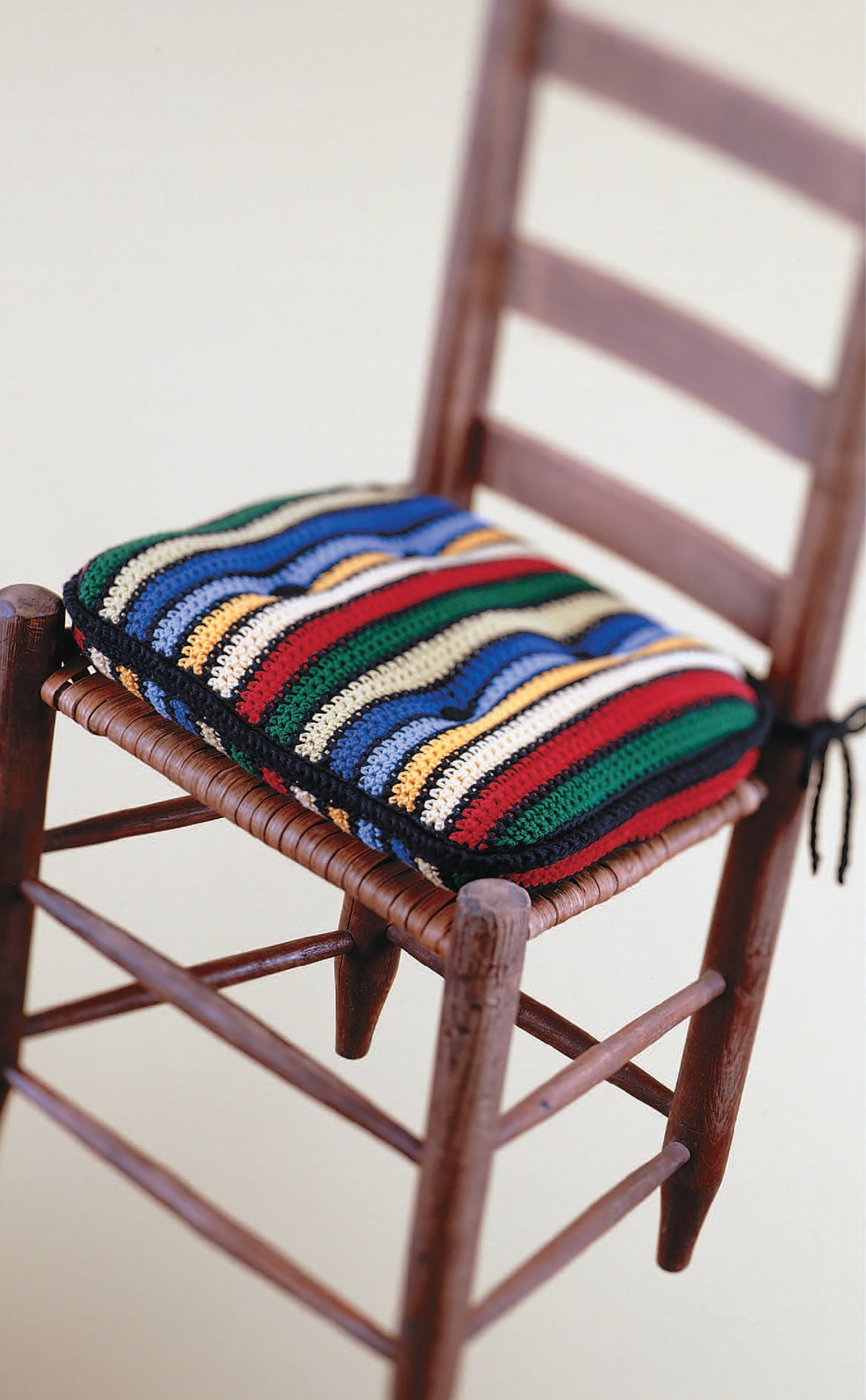 The Little Kitchen Helpers cushion by Leisure Arts features colorful, striped crochet inspired by handmade dishcloths, adding an eye-catching accent to a wooden ladder-back chair with a woven seat.