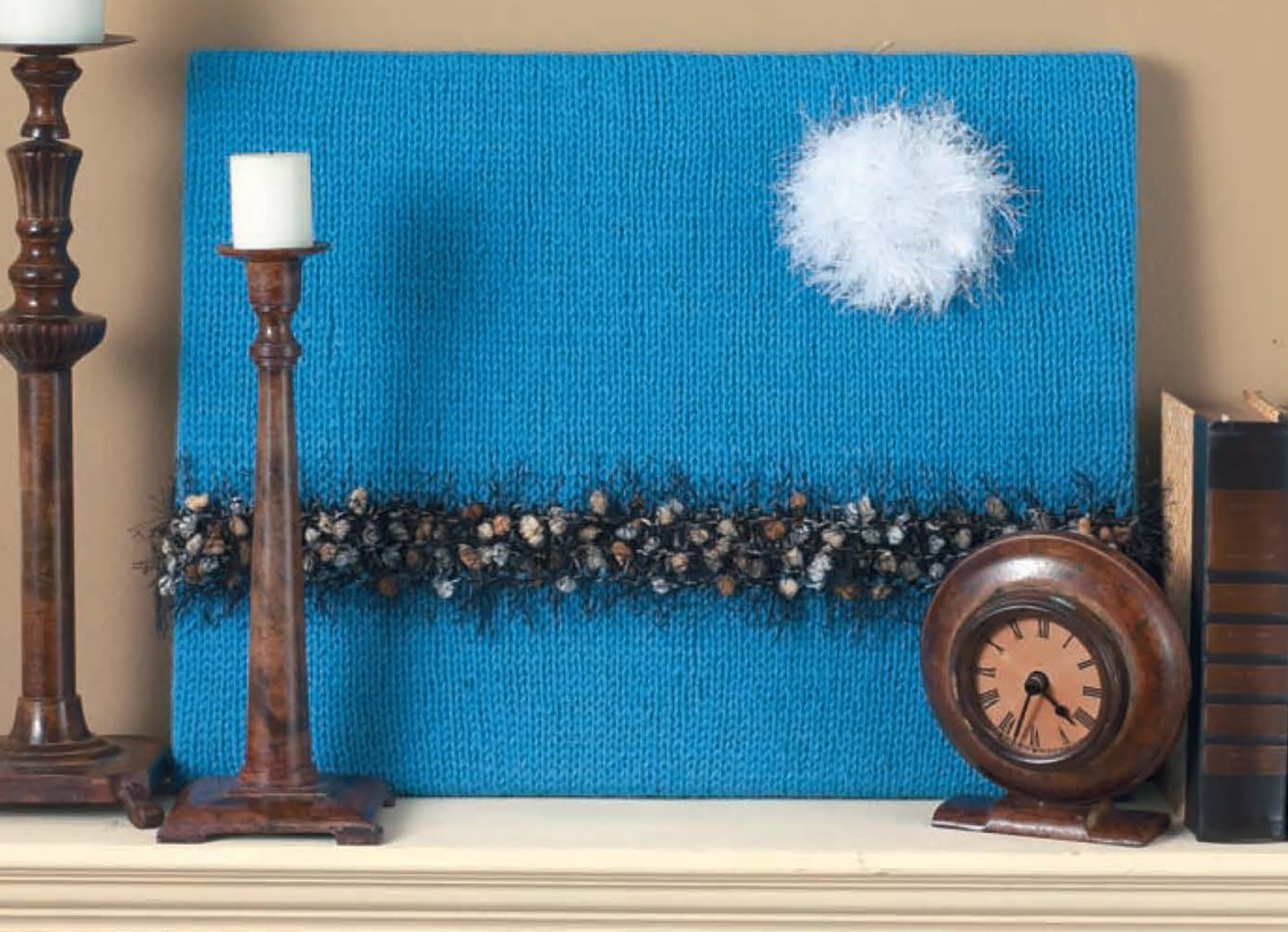 The Modern Punch Needle by Leisure Arts features blue textured artwork with a white fluffy circle and a dark fuzzy band—displayed on a shelf with wooden candlesticks, clock, and books—perfect inspiration for beginner projects.