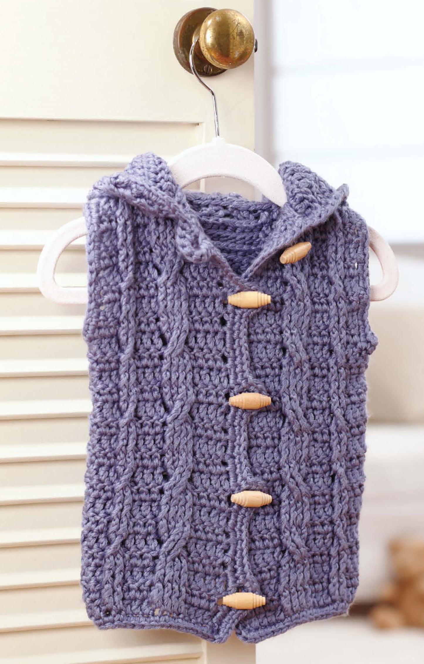 The Sophisticated Babies & Toddlers—6 Crochet Designs Digital Download by Leisure Arts features patterns like a hand-knitted, sleeveless lavender hooded vest with wooden buttons, perfect for cozy style, shown on a hanger over a brass door handle.