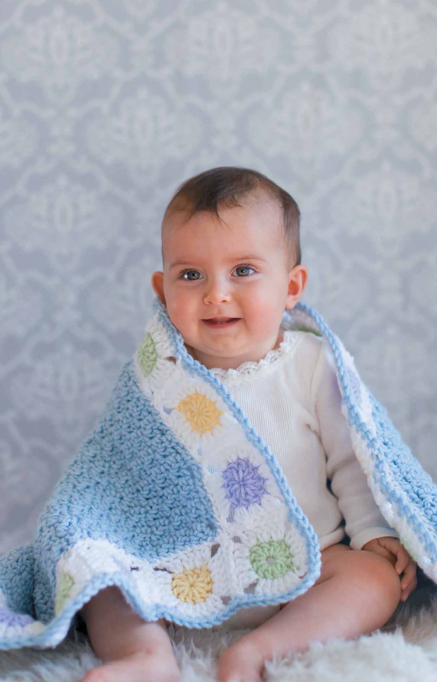 A blue-eyed baby sits on a soft surface, wrapped in Leisure Arts' Baby Afghans—Cuddly Wraps for Babies & Toddlers!, a pastel blue and white crocheted blanket with beginner-friendly patterns and colorful flower motifs.