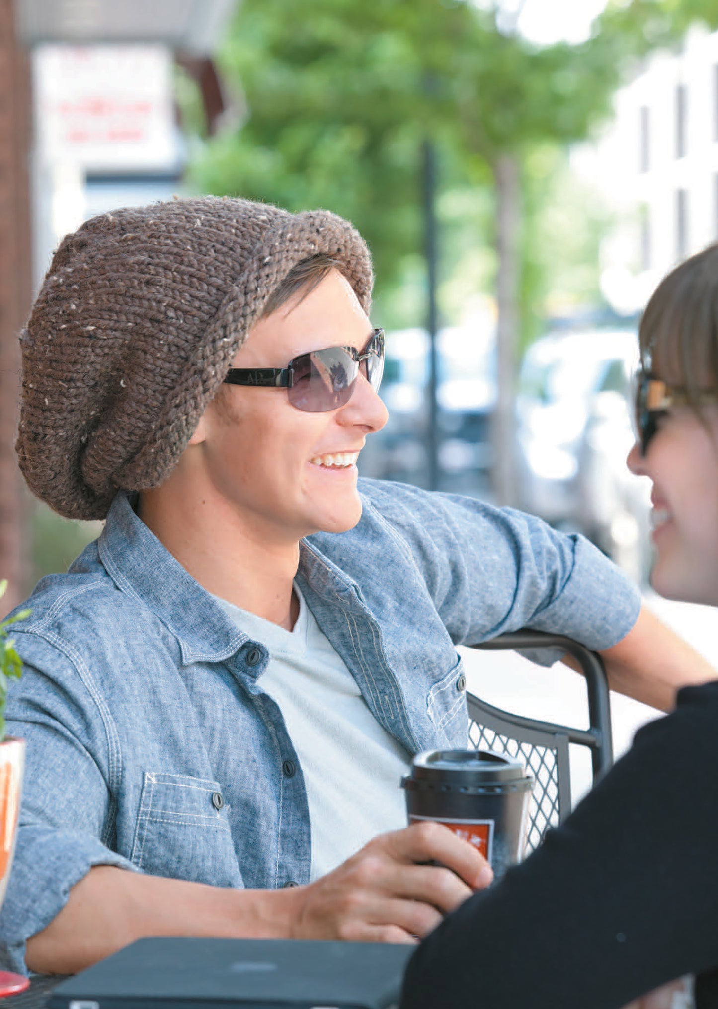 A person in sunglasses and a denim shirt smiles with a coffee cup, wearing a beanie made from the "Knit Celebrity Slouchy Beanies for the Family Book 2 Digital Download" by Leisure Arts, at an outdoor café with trees and parked cars behind.