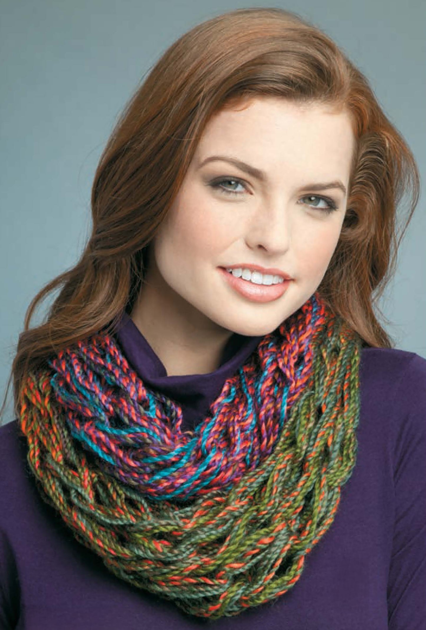 A woman with long auburn hair and light makeup smiles at the camera in a purple top, wearing a chunky multicolored cowl made using "Learn to Arm Knit" by Leisure Arts, against a gray background.